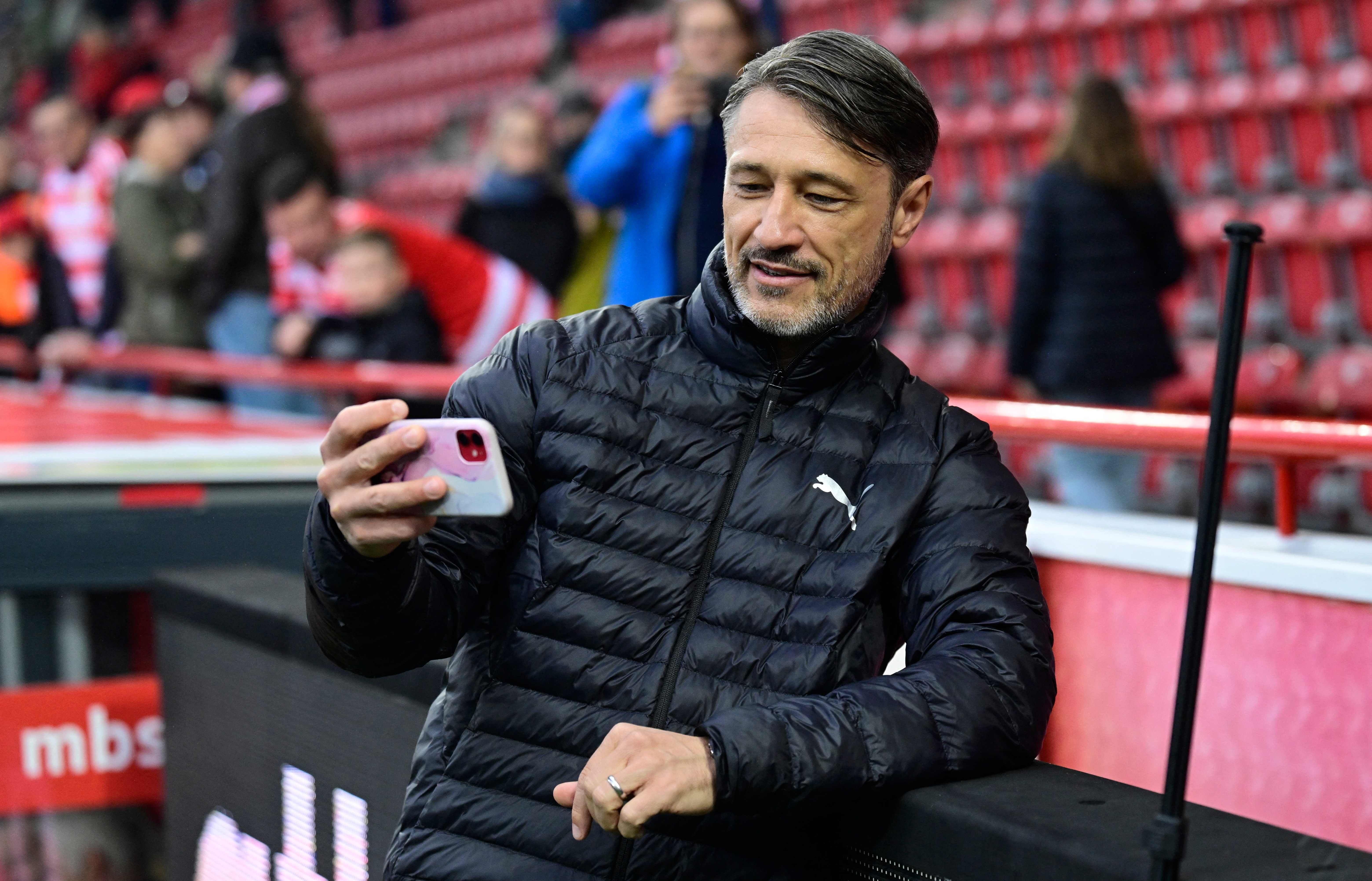 Niko Kovac steht in einer schwarzen Daunenjacke im Stadion an der Bandenwerbung und blickt schmunzelnd auf sein in der Hand gehaltenes Smartphone.