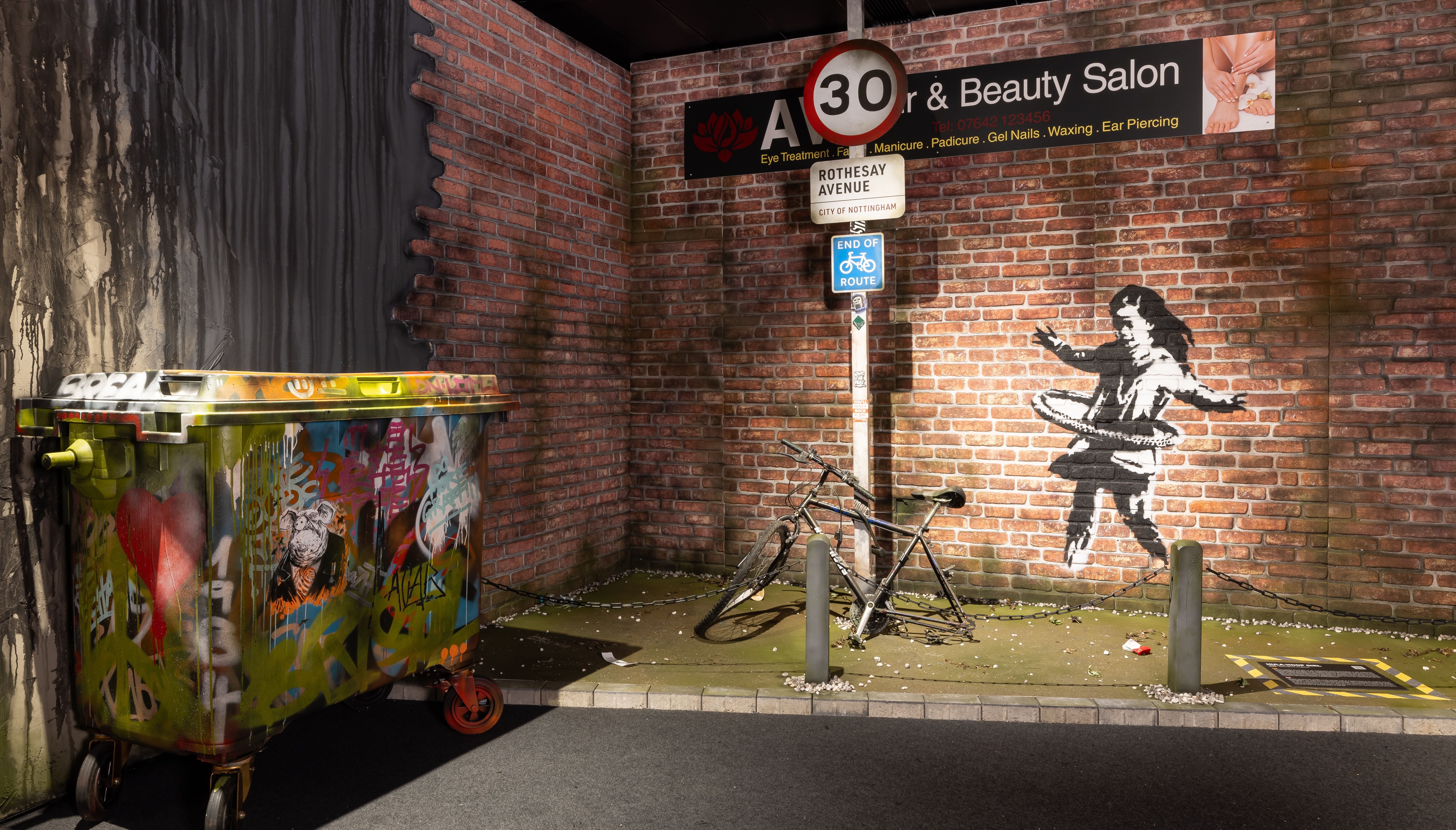 Banksy-Graffiti eines Mädchens mit Fahrradreifen, daneben ein bunt besprühter Müllcontainer und ein kaputtes Fahrrad.