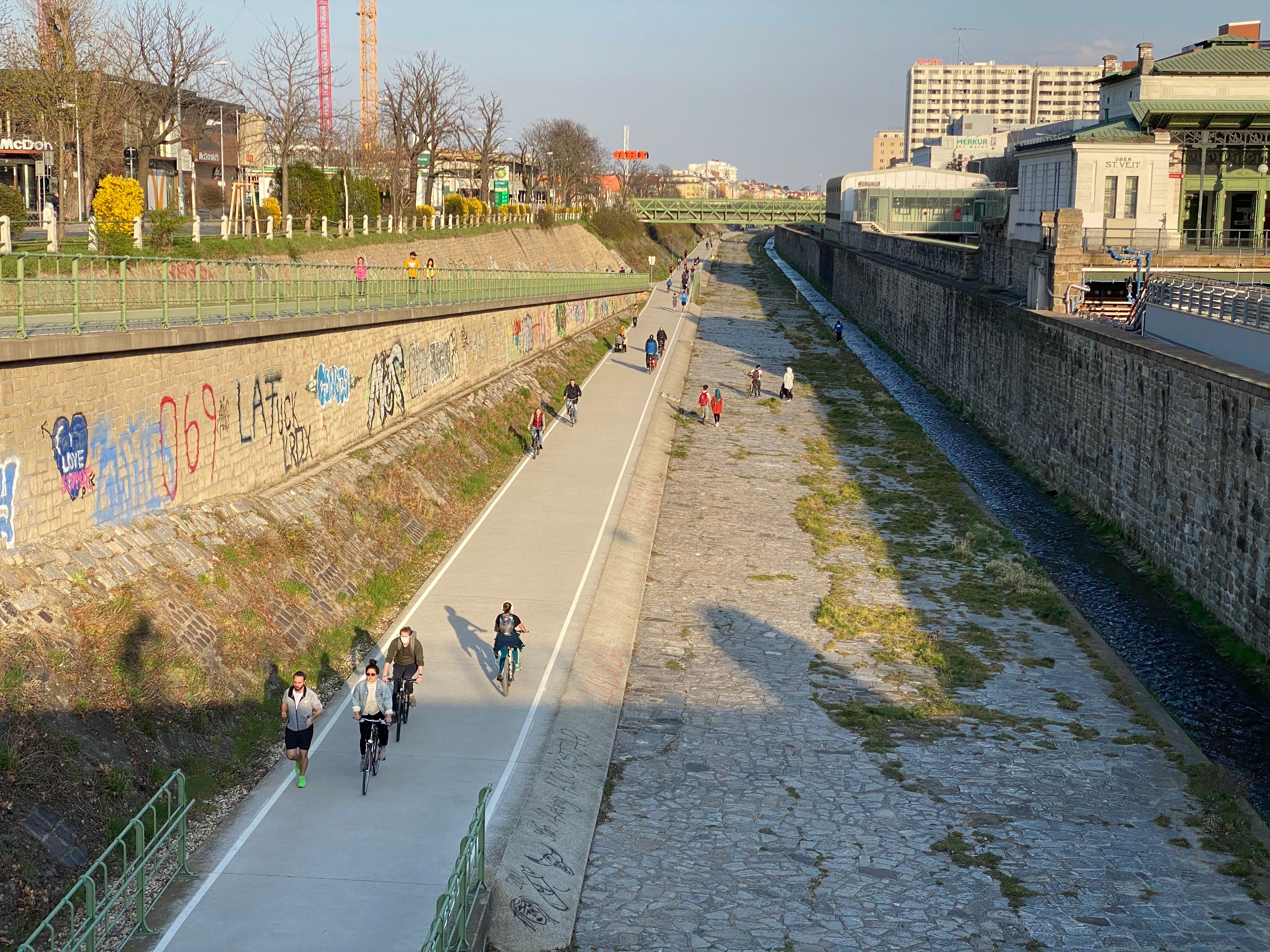 Menschen gehen und fahren Fahrrad auf einem Weg entlang eines betonierten Kanals mit Hochwasserschutzmauer.