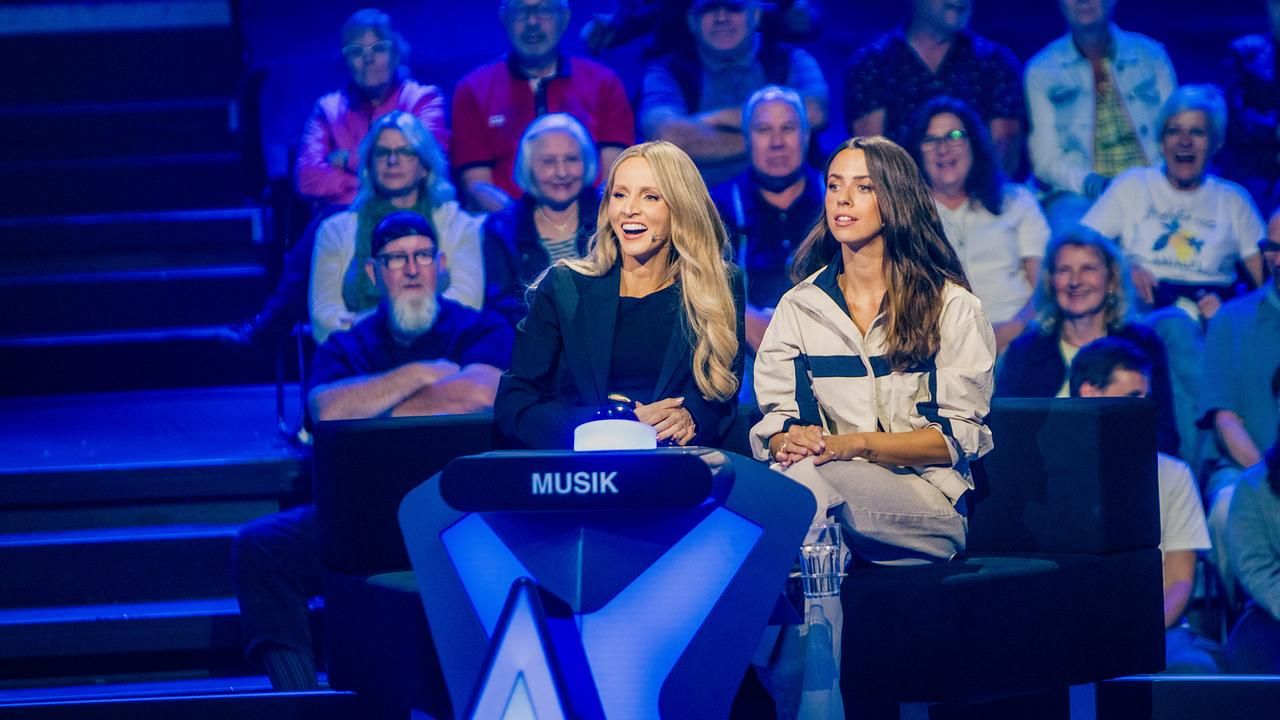 Zwei Frauen sitzen an einem Musik-Quizpult, umgeben von einem Publikum in einem Studio.