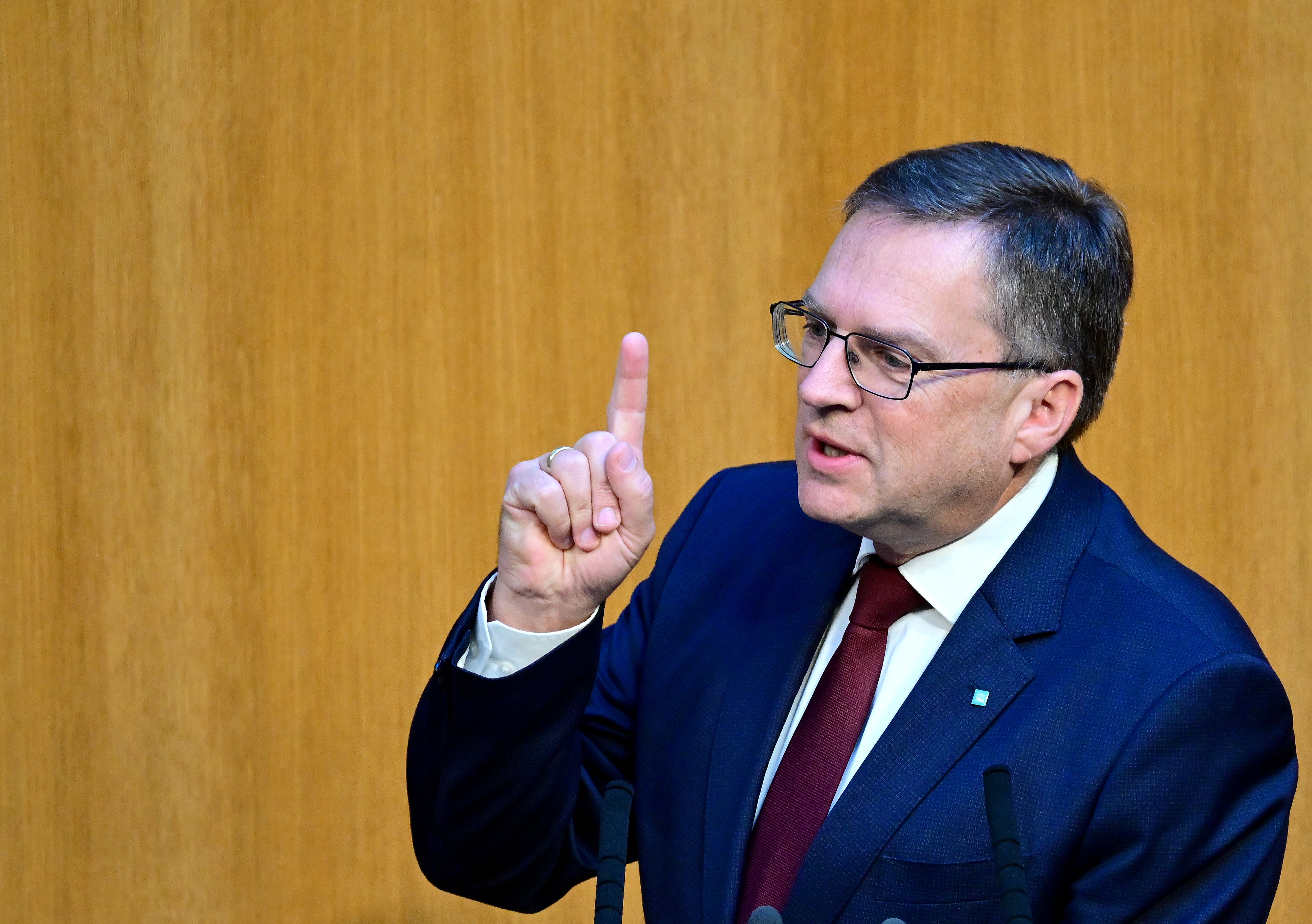 Ein Mann im Anzug spricht und hebt den Zeigefinger, Holzwand im Hintergrund.