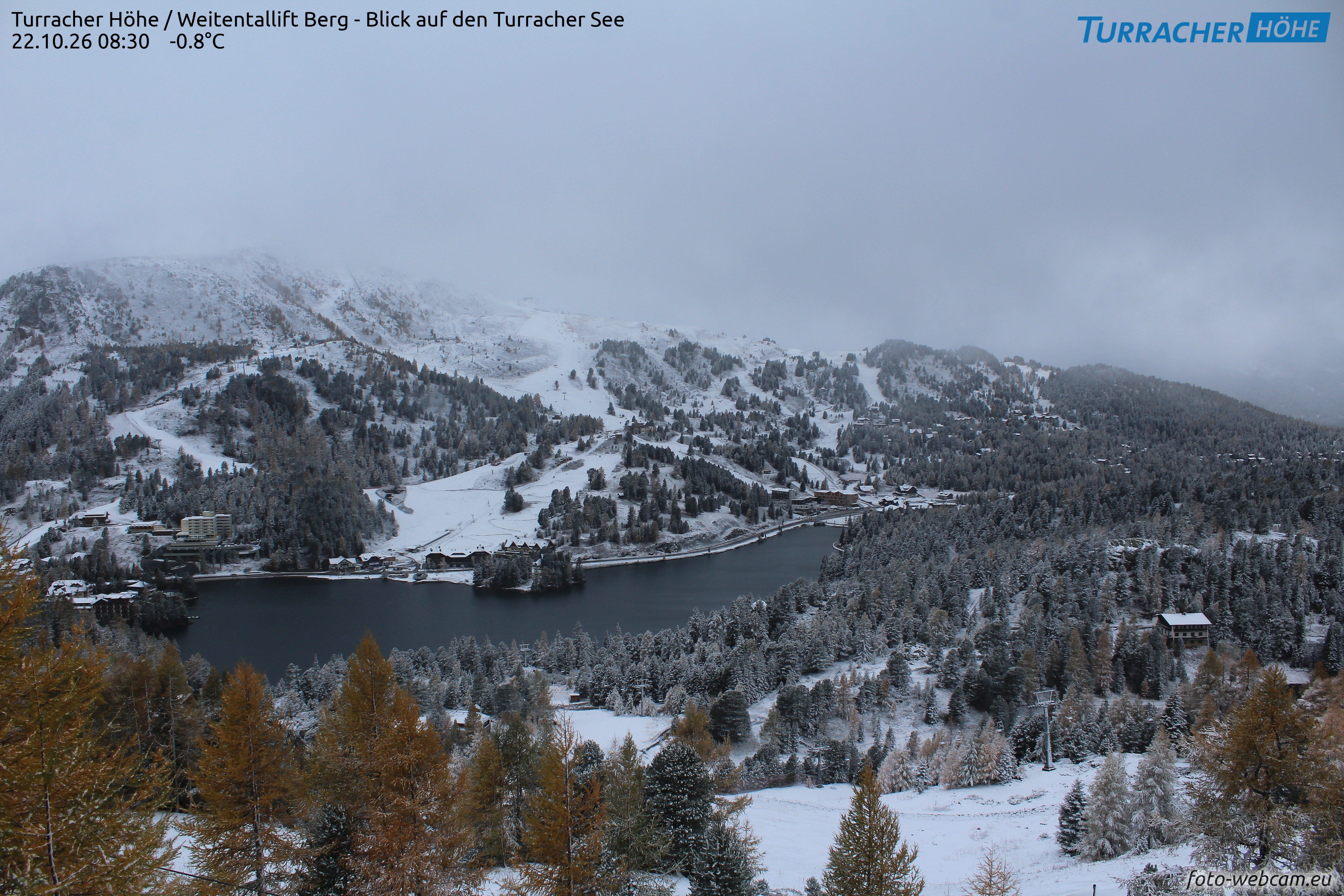 Blick auf den verschneiten Turracher See und umliegende Berge unter bewölktem Himmel.