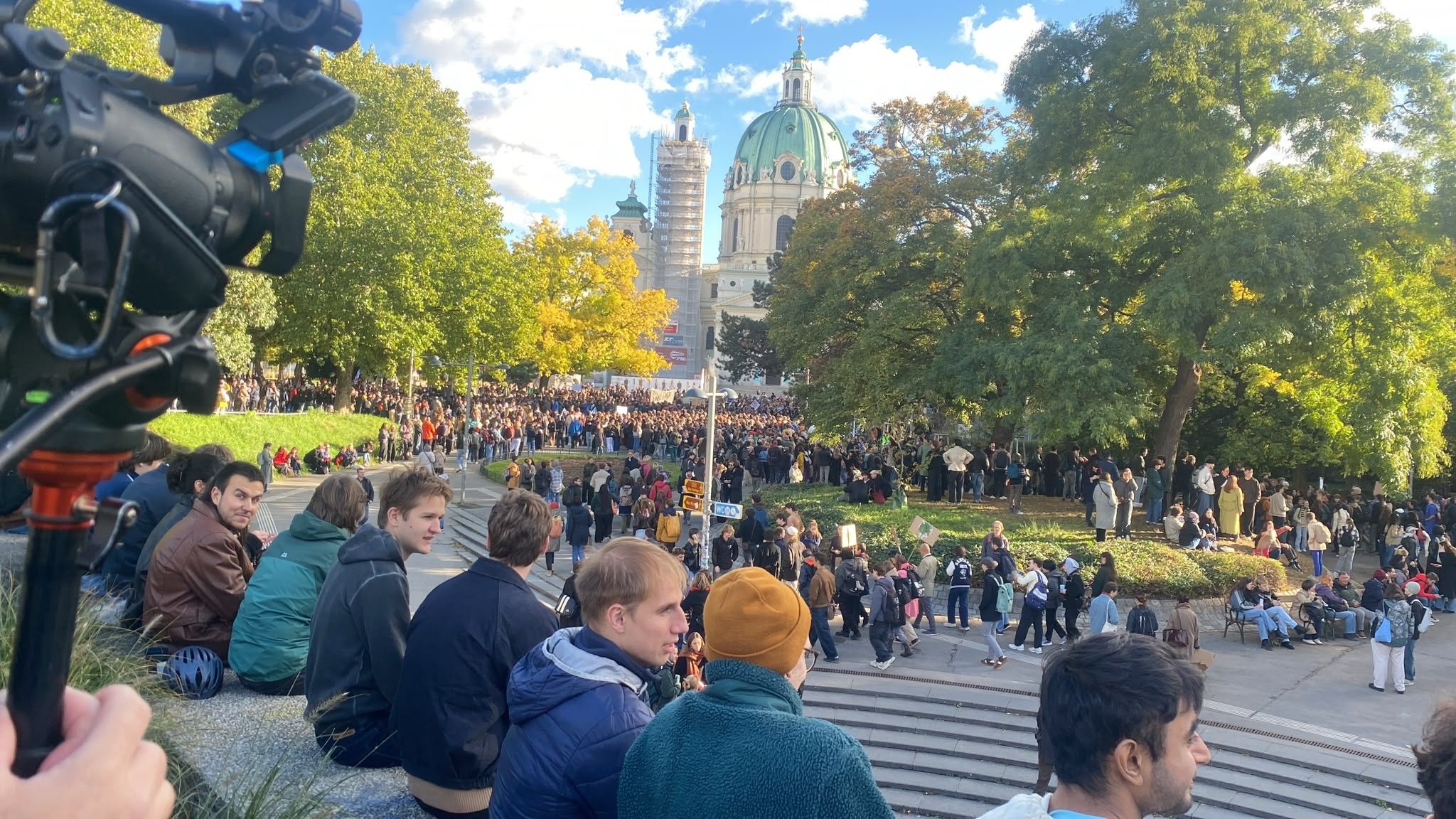 Menschenmenge auf einer Demo im Park, viele sitzen auf Stufen, Kamera links, Kirche im Hintergrund.