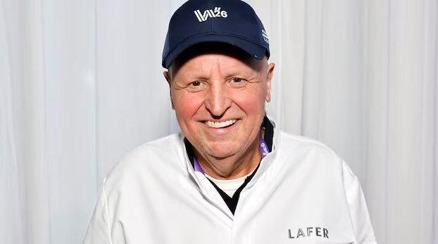 Ein lächelnder Mann in weißer Jacke und blauer Kappe steht vor weißem Hintergrund.