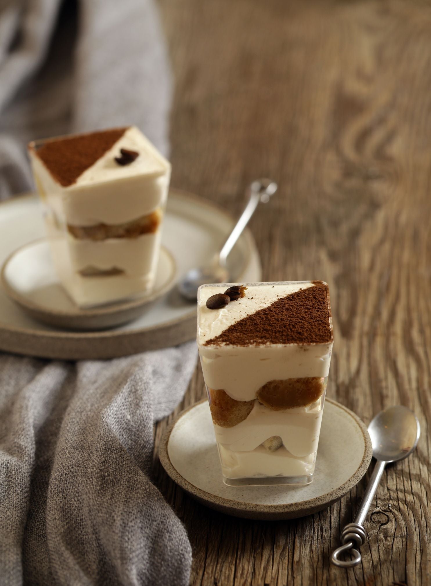 Zwei Gläser Tiramisu auf Tellern mit Löffeln, dekorativ angerichtet auf Holztisch.