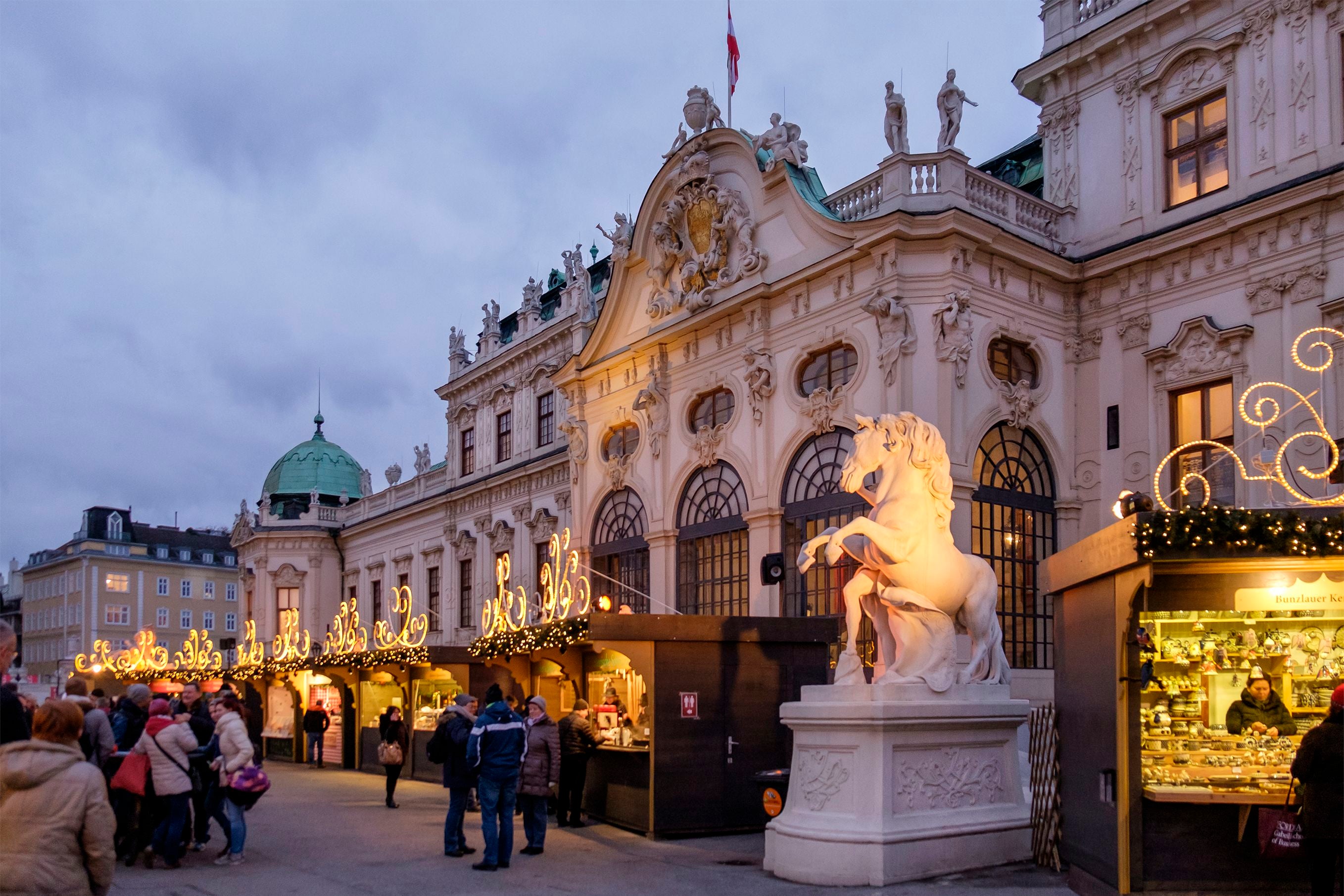 Menschen besuchen einen festlich beleuchteten Weihnachtsmarkt vor dem Belvedere in Wien.