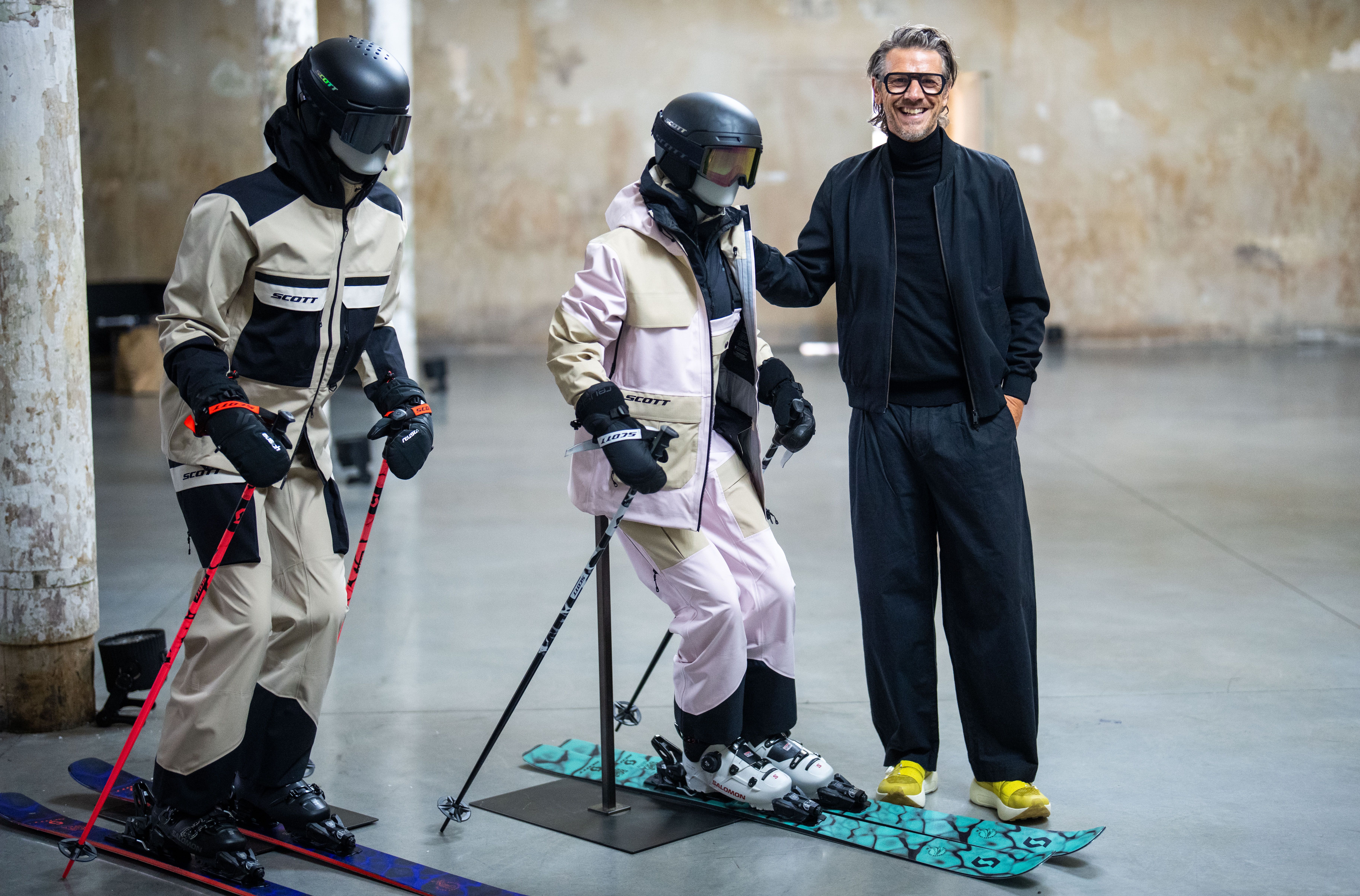 Ein Mann steht neben zwei Skimodellen in Skianzügen mit Helmen und Skibrillen in einer Halle.