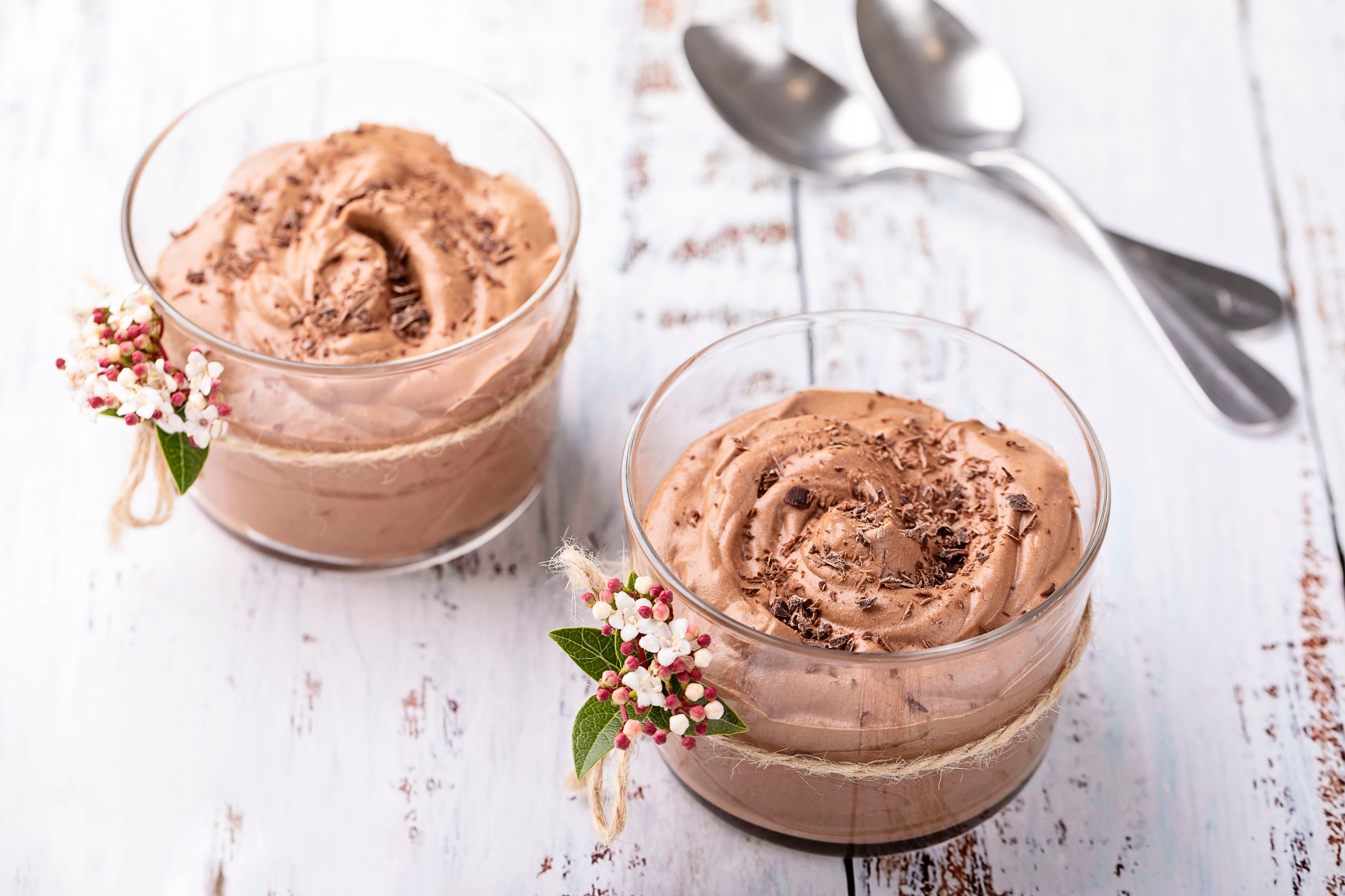 Zwei Gläser Schokoladenmousse mit Deko auf weißem Holztisch, dahinter zwei Löffel.