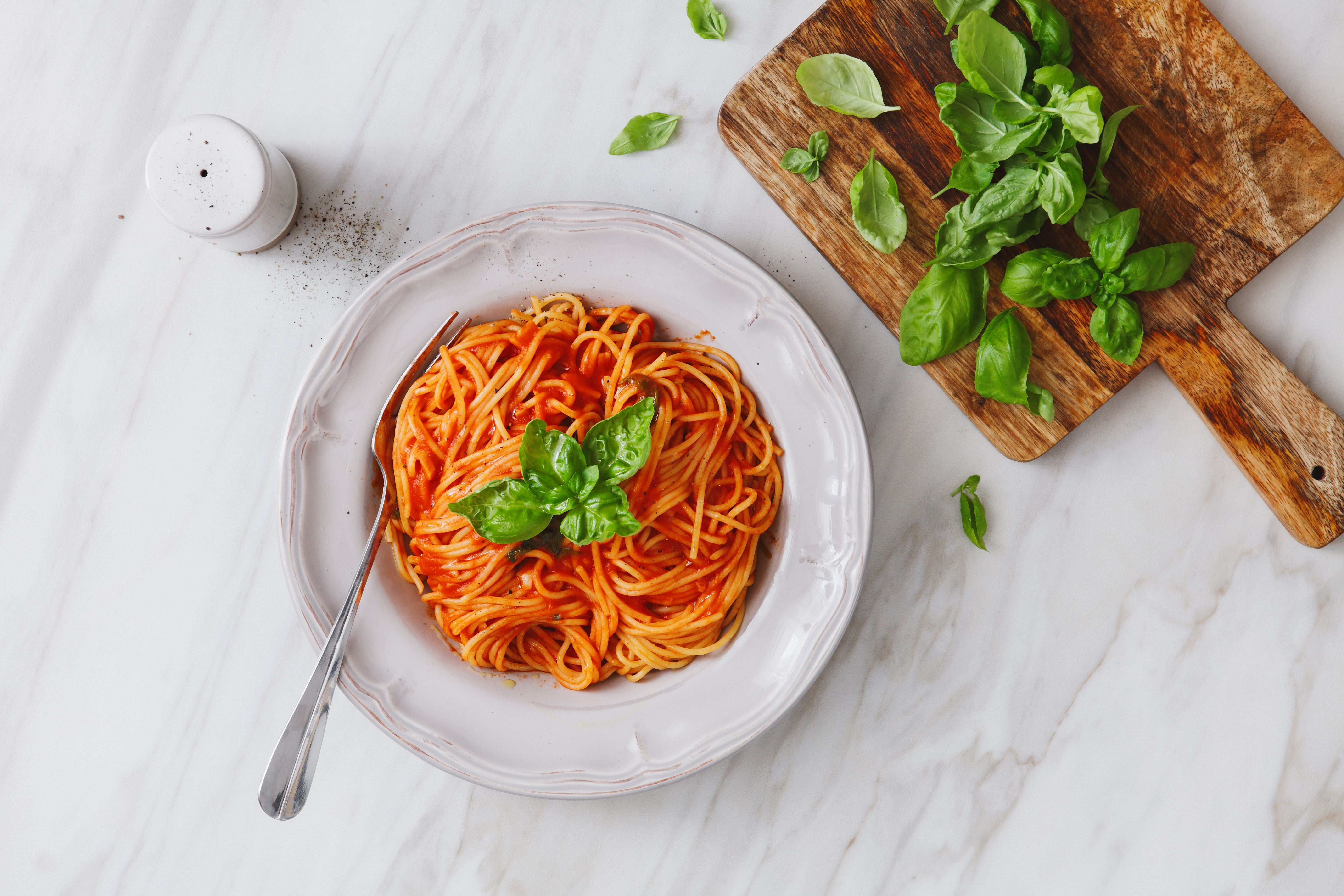 Teller Spaghetti mit Tomatensauce und Basilikum, daneben frischer Basilikum auf einem Holzbrett.