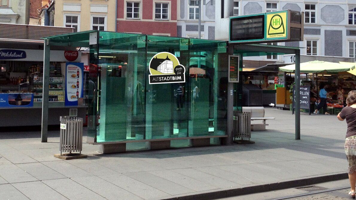 Gläserne Straßenbahnhaltestelle der Altstadtbim in Graz mit Sitzbänken und Marktständen im Hintergrund.