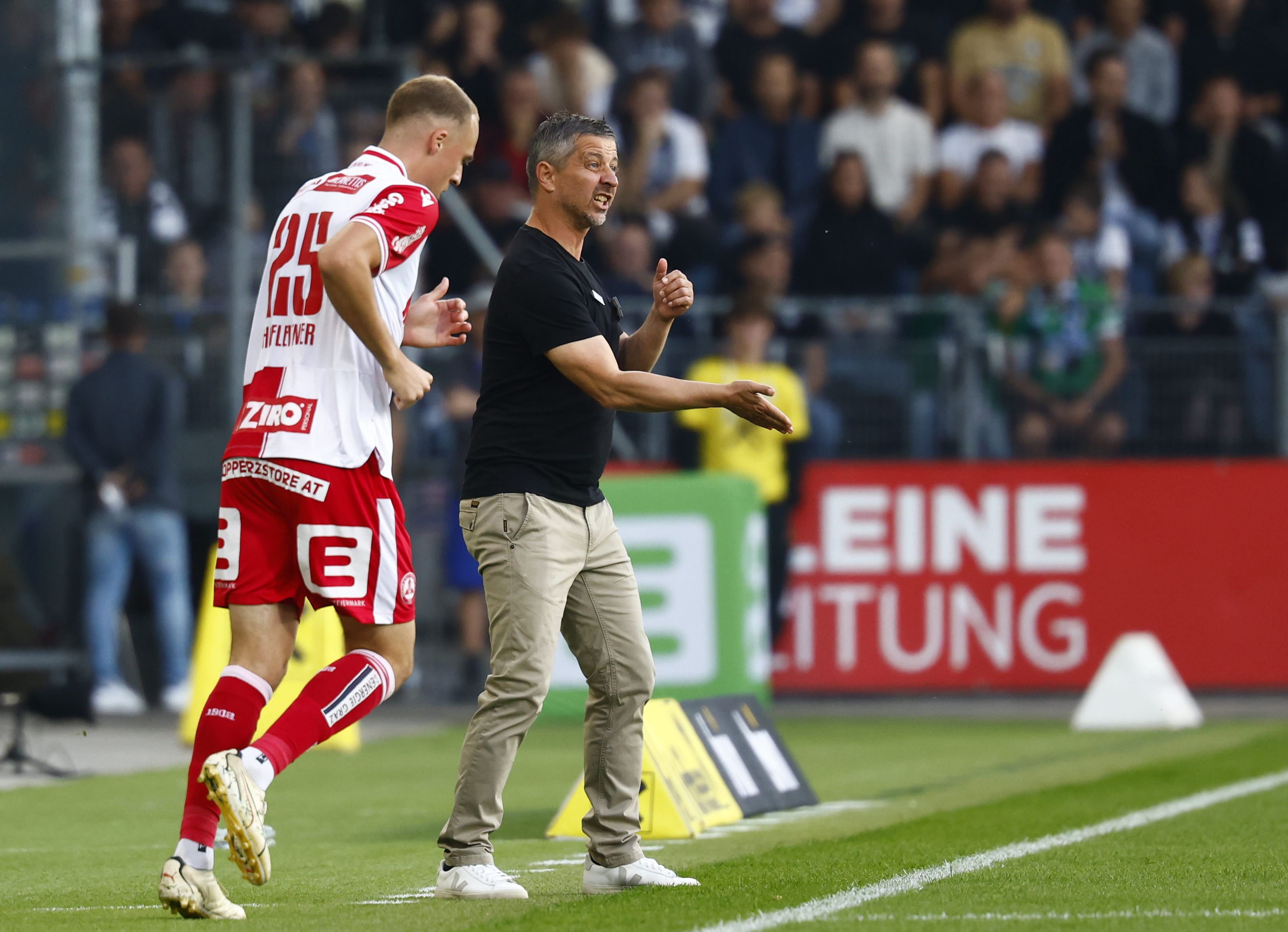 GAK-Trainer Gerald Scheiblehner gibt im schwarzen T-Shirt an der Seitenlinie energisch Anweisungen an einen vorbeilaufenden Spieler im rot-weißen Trikot.