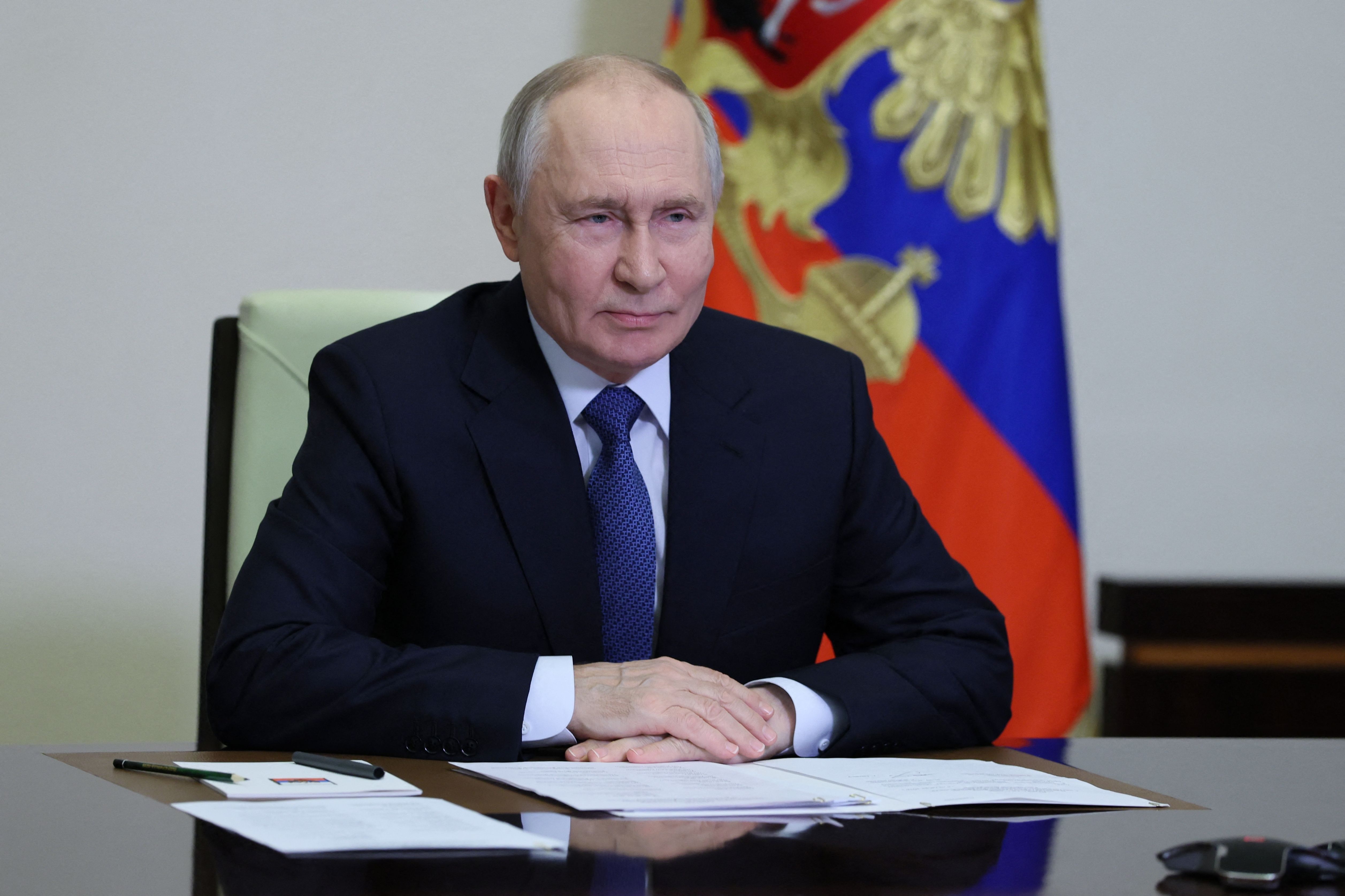 Ein Mann im Anzug sitzt an einem Tisch mit Papieren, im Hintergrund eine russische Flagge.