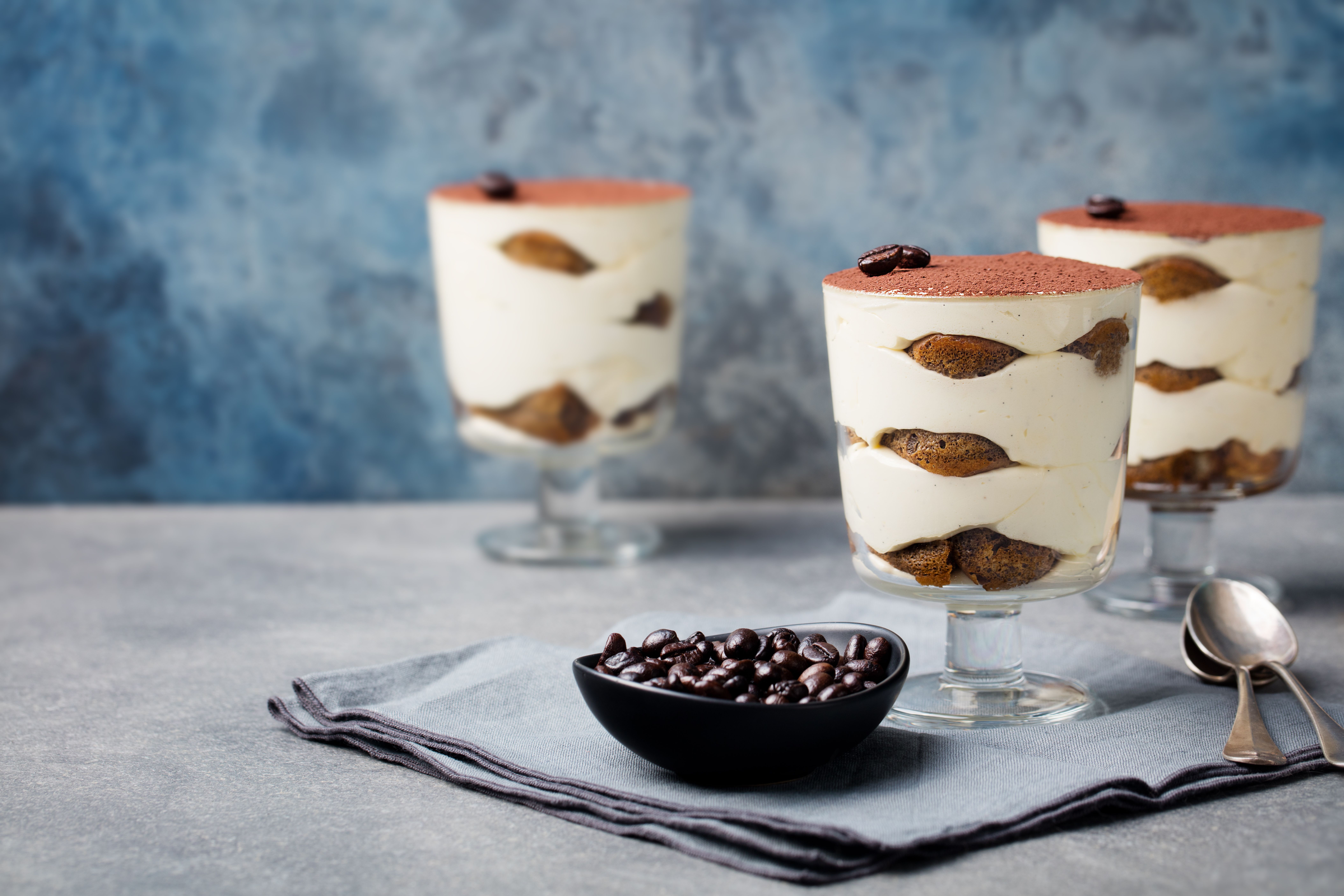 Drei Gläser Tiramisu mit Löffelbiskuit, davor eine Schale Kaffeebohnen und zwei Löffel.