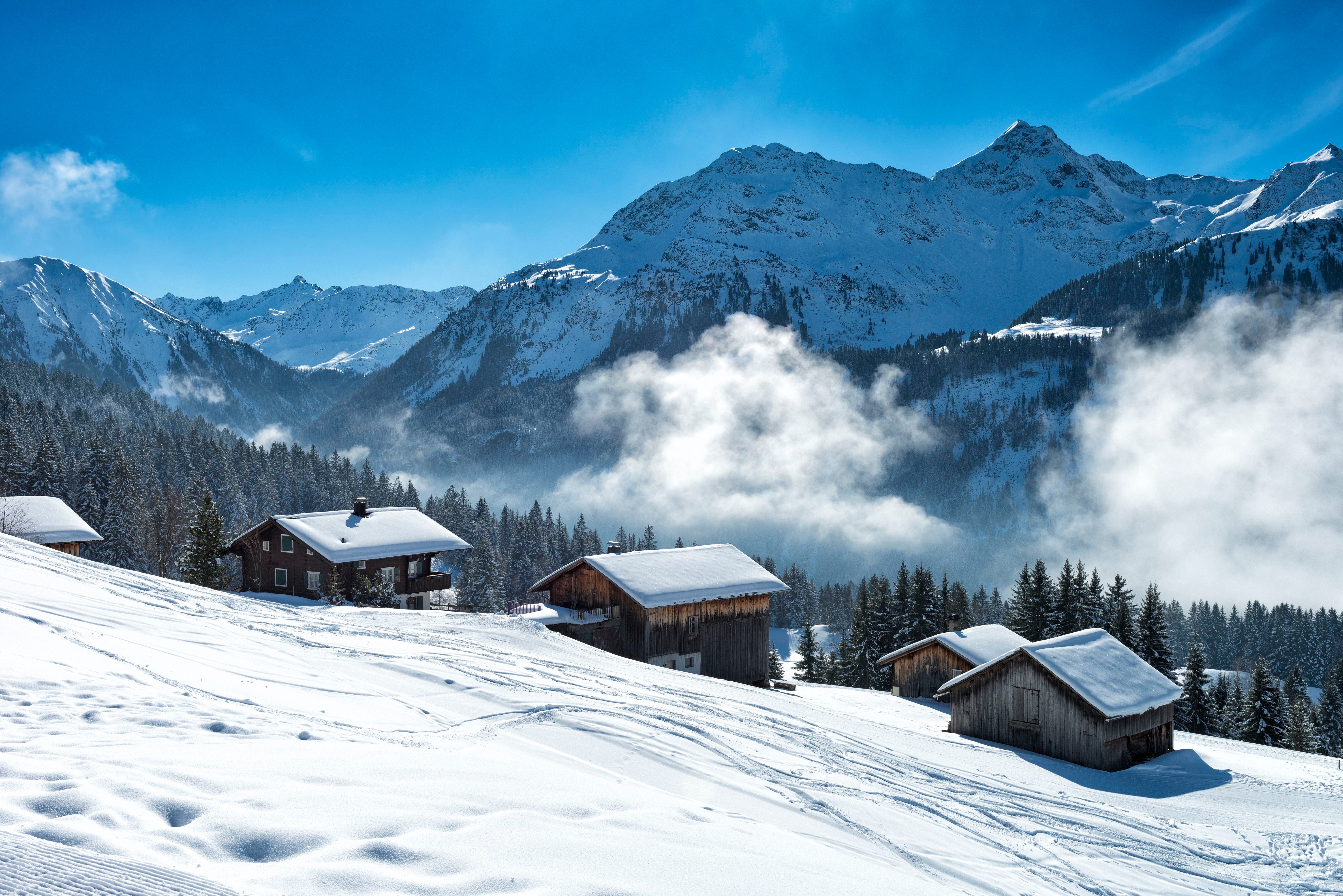 Verschneite Berghütten vor Alpenpanorama mit Nebelschwaden und Wald in Vorarlberg, Österreich.