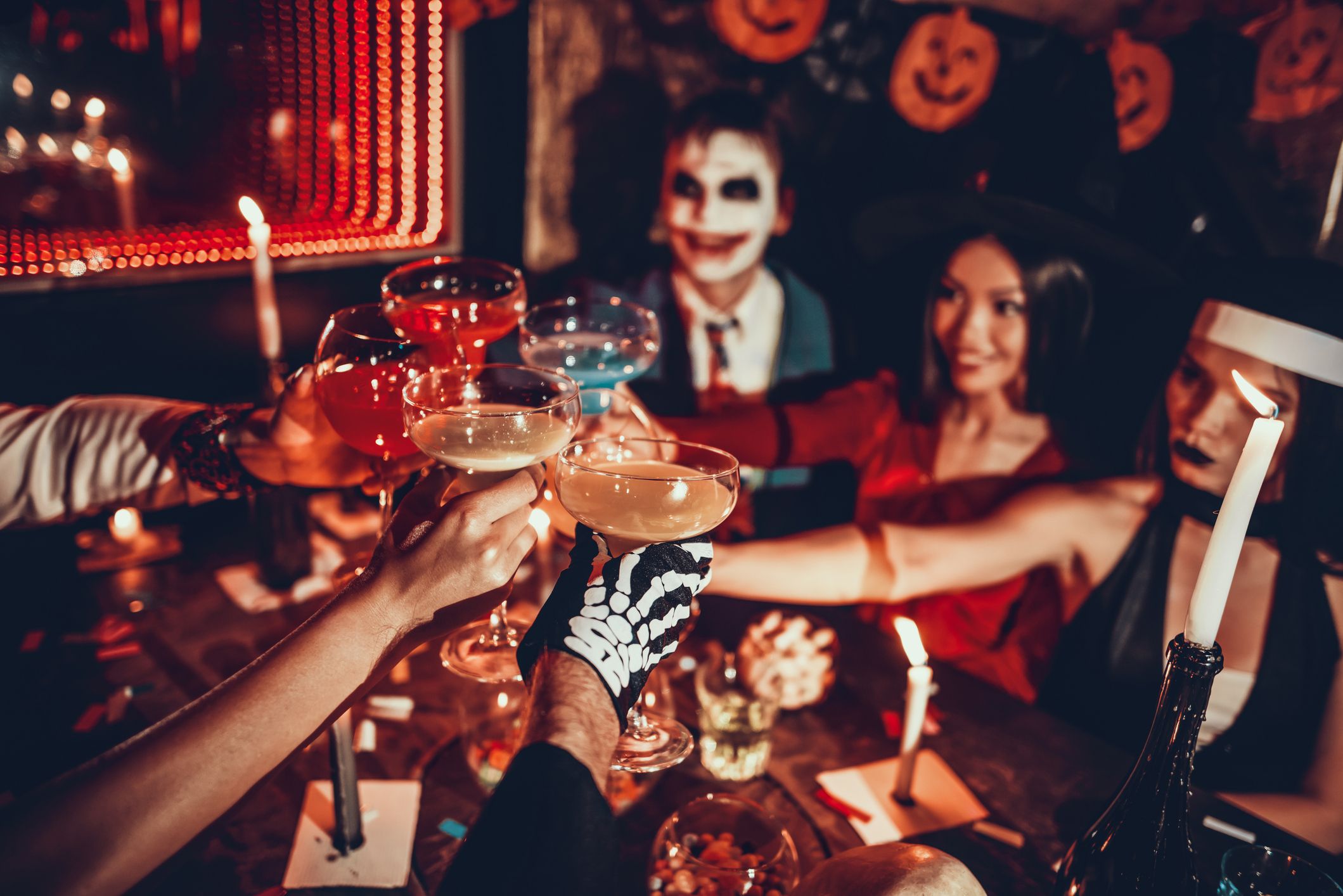 Junge Leute in Halloween-Kostümen stoßen mit Cocktails an einem festlich dekorierten Tisch an.