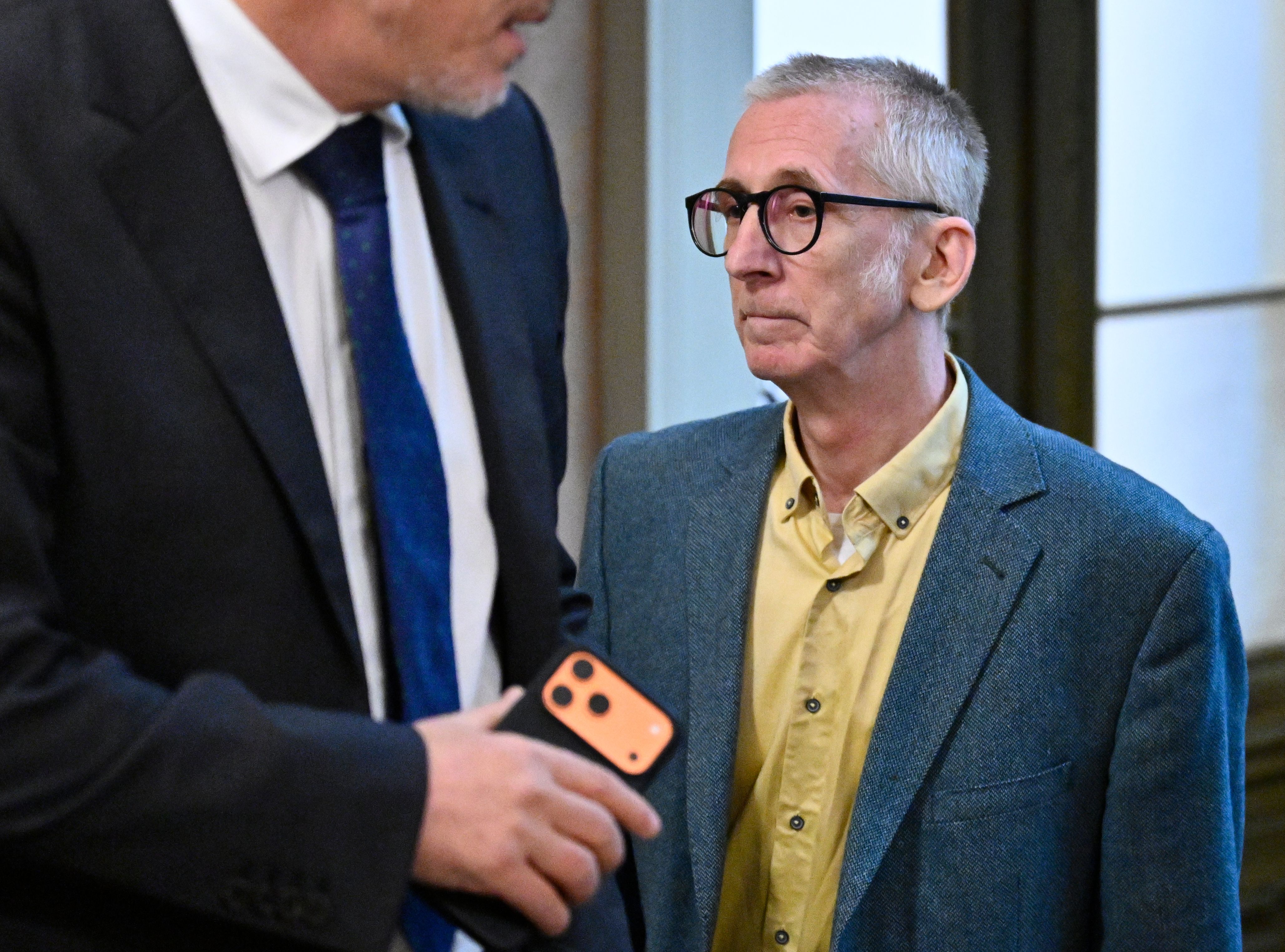 Ein Mann mit Brille und gelbem Hemd steht neben einer Person im Anzug in einem Gerichtssaal.