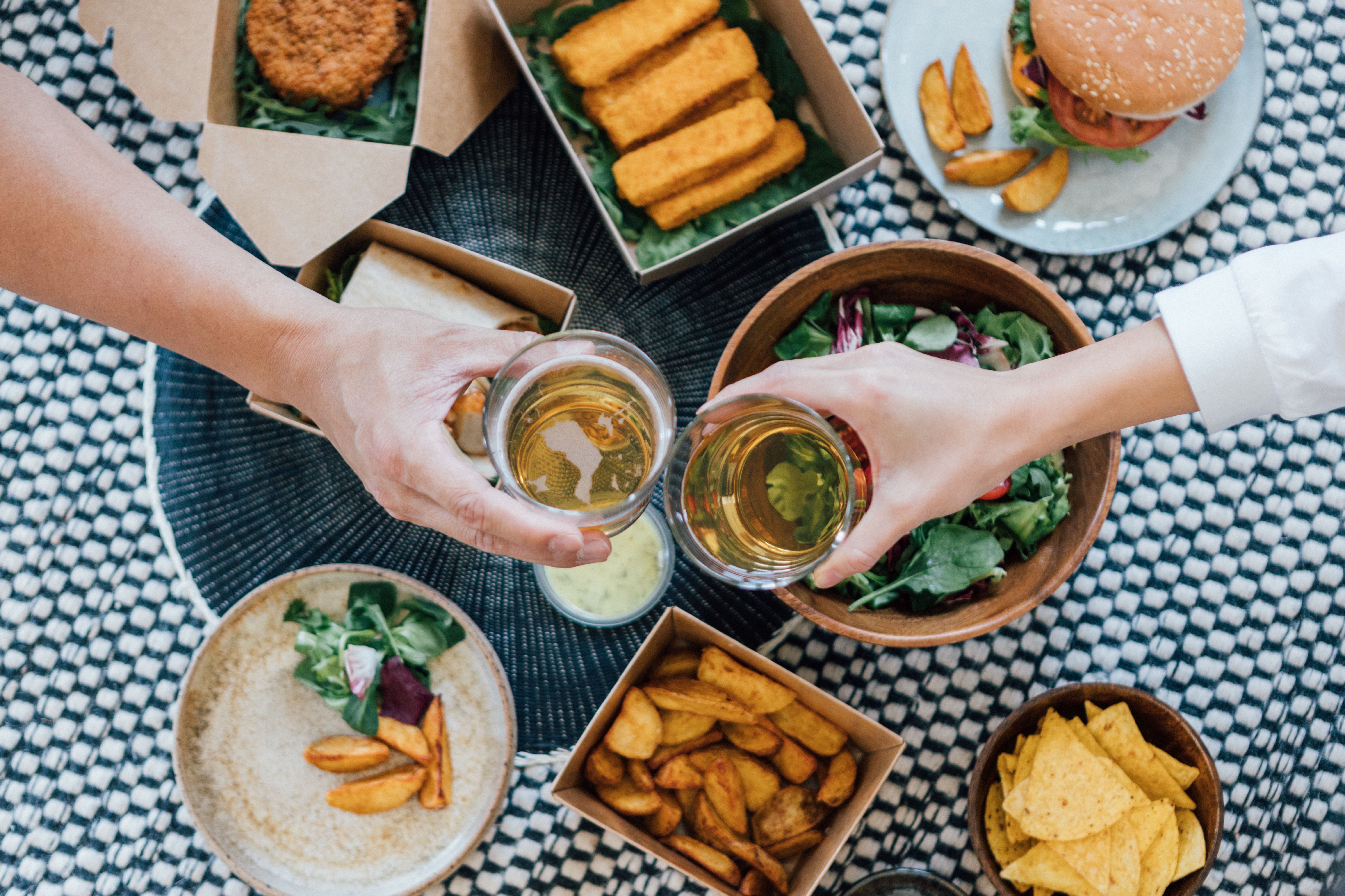 Zwei Personen stoßen mit Bier an, umgeben von Fast Food wie Burger, Pommes und Salat.