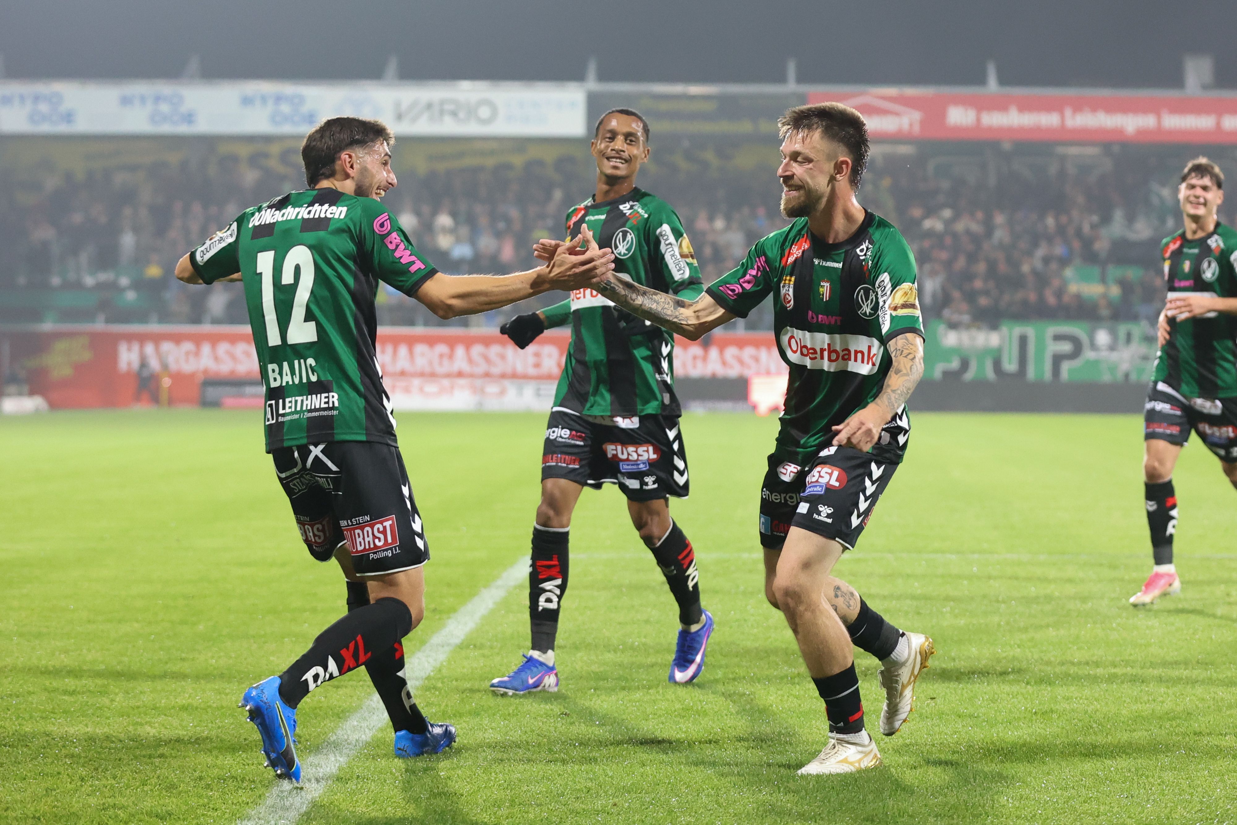 Fünf Fußballer der SV Ried jubeln gemeinsam in ihren grün-schwarzen Trikots auf dem Rasen.