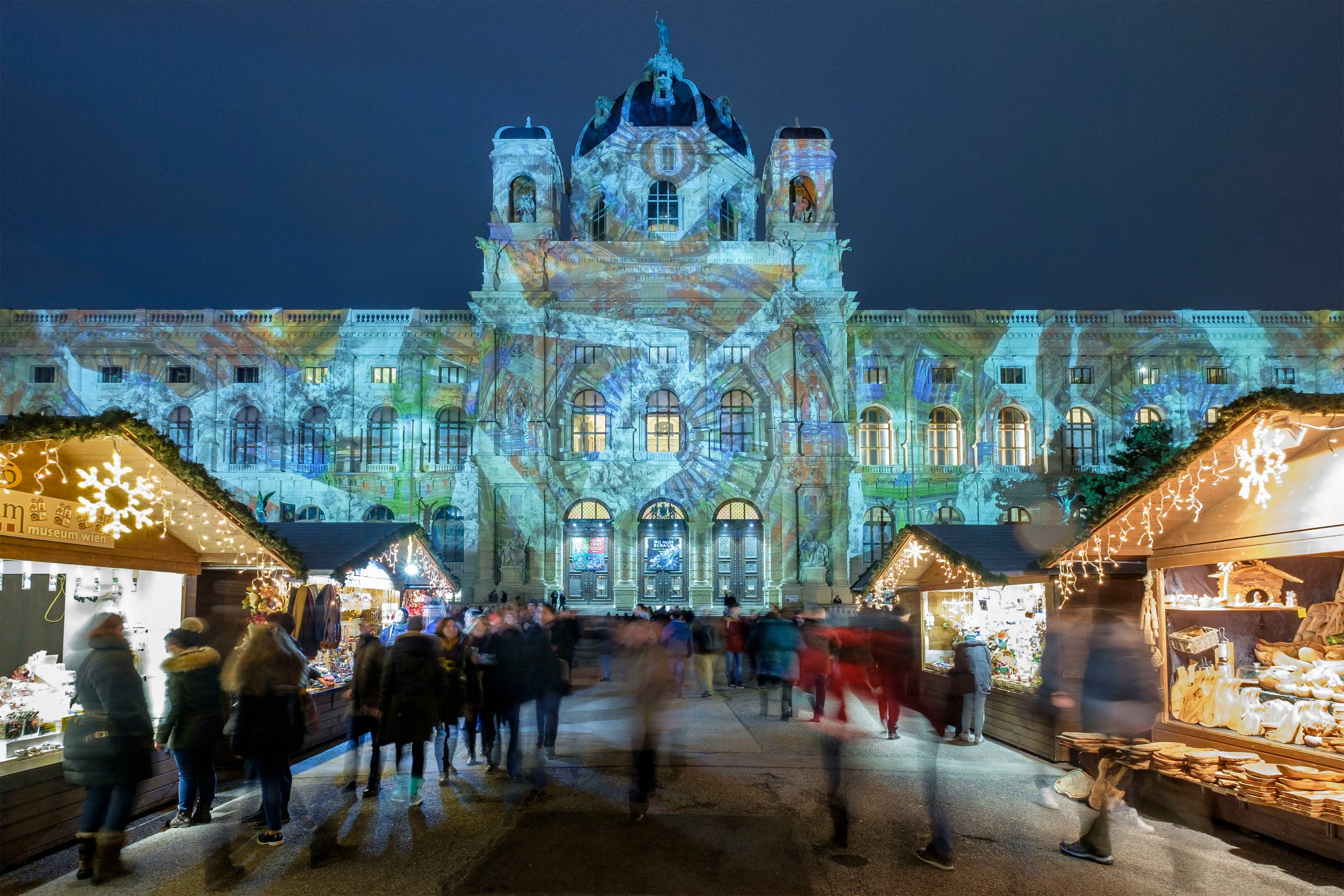 Weihnachtsmarkt mit Lichtprojektionen am Naturhistorischen Museum in Wien, Menschen schlendern vorbei.