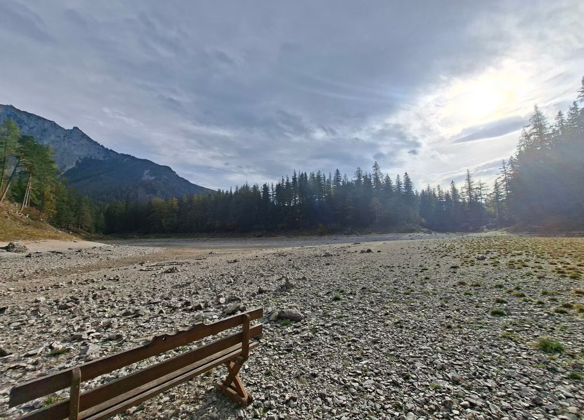 Holzbank steht auf steinigem, ausgetrocknetem Seebett vor bewaldeten Bergen und bewölktem Himmel.