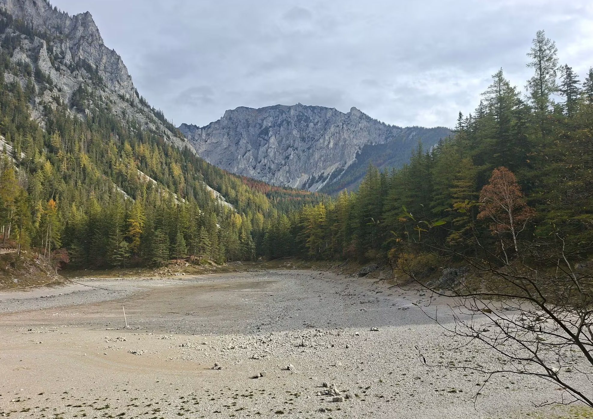 Ausgetrockneter Grüner See, umgeben von bewaldeten Bergen und Felsen unter bewölktem Himmel.
