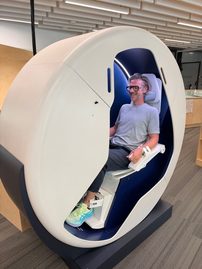 Ein Mann sitzt lächelnd in einer futuristischen Kapsel im Inneren eines modernen Gebäudes.