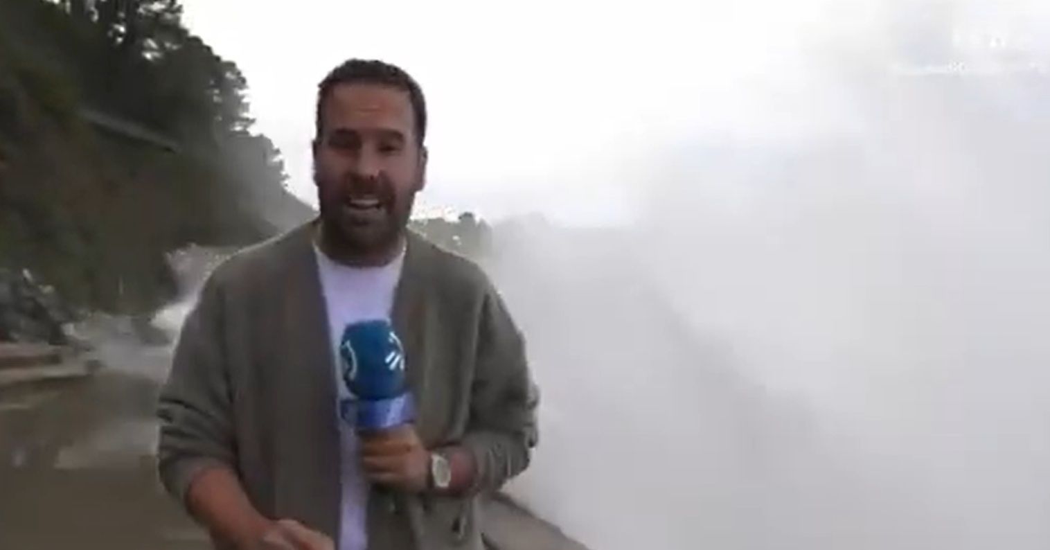 Reporter steht am Strand, während eine riesige Welle im Hintergrund aufschlägt.