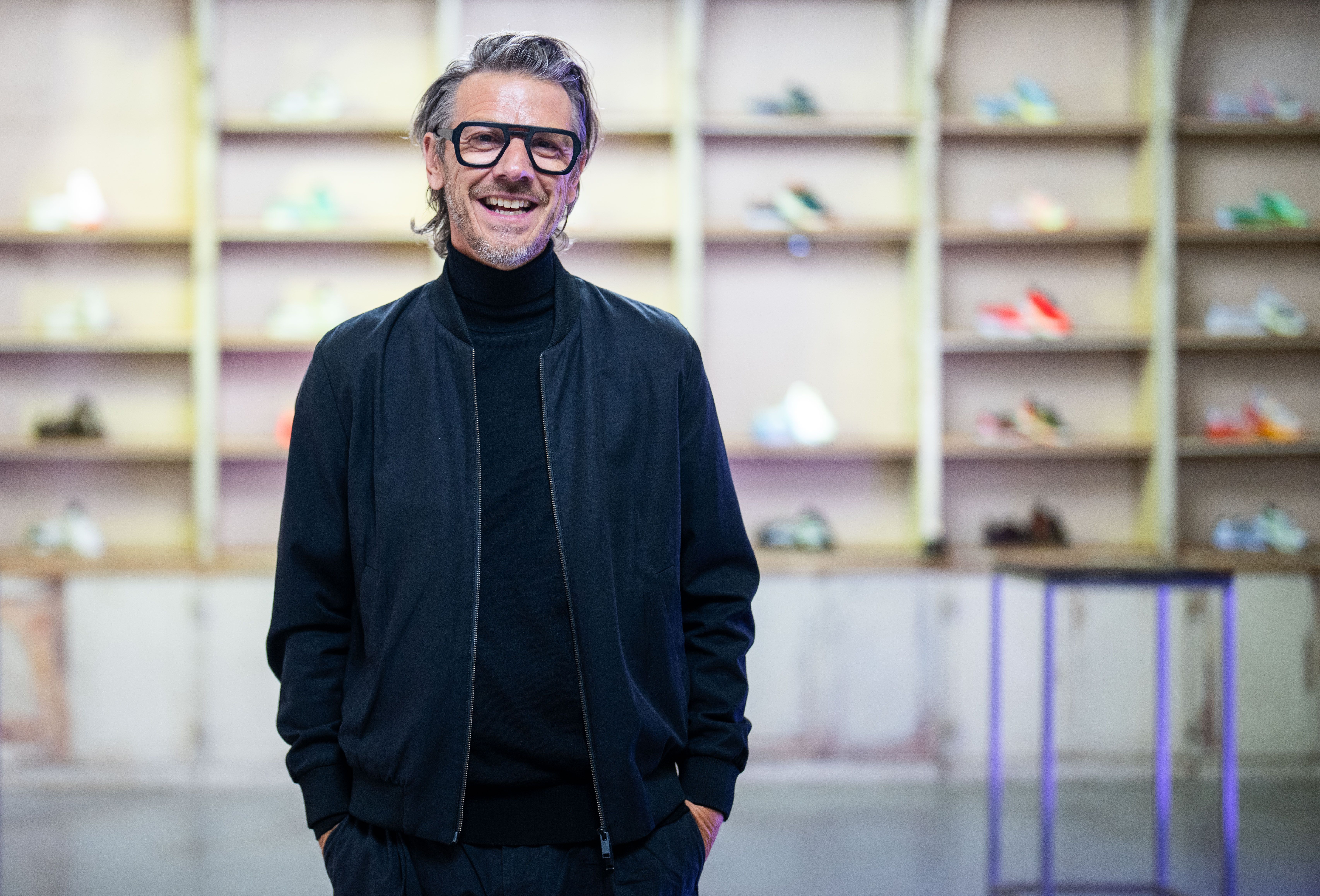 Ein Mann mit Brille und dunkler Kleidung steht lächelnd vor Regalen mit Sportschuhen.
