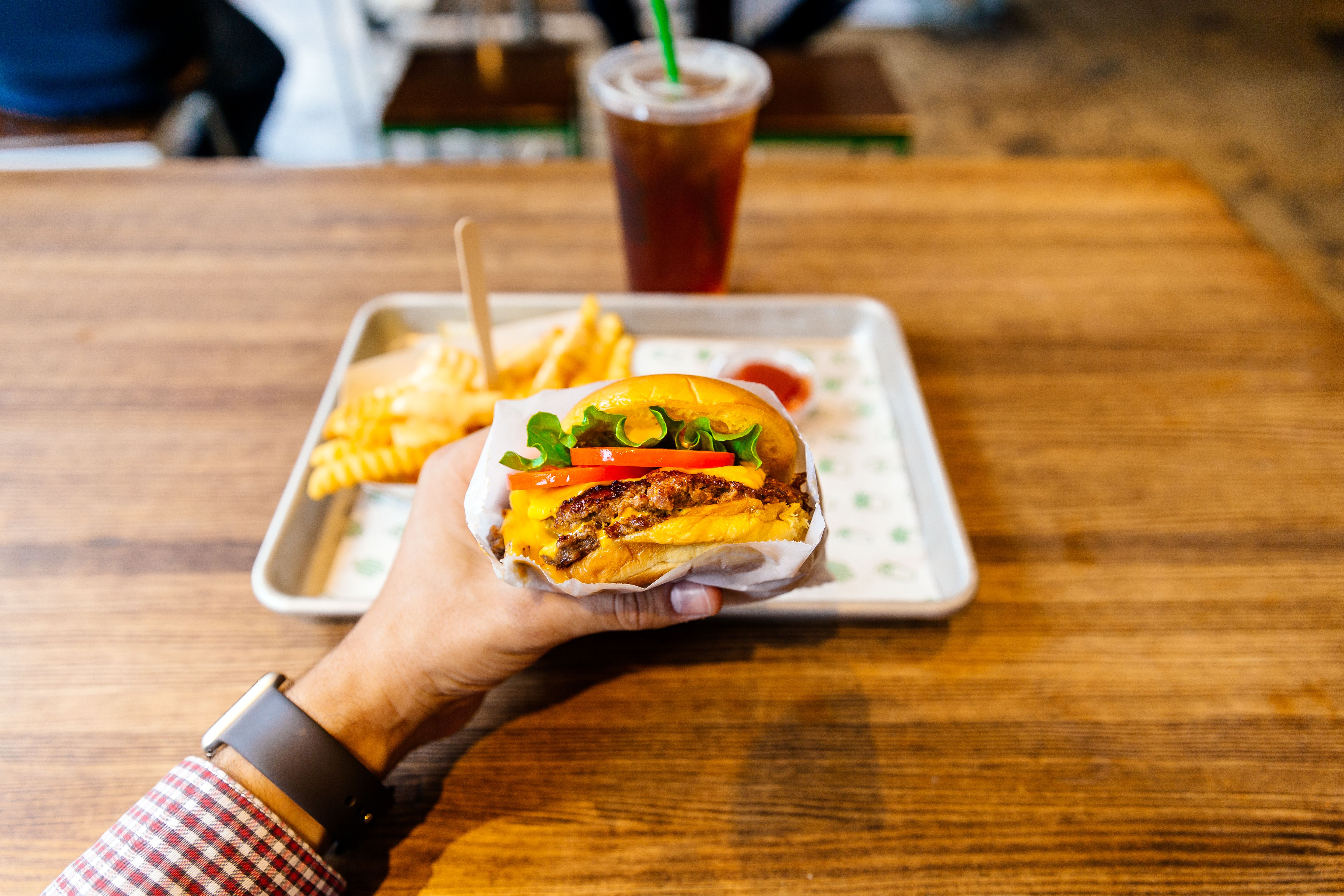 Eine Hand hält einen Cheeseburger; im Hintergrund Pommes, Ketchup und ein Getränk auf dem Tisch.