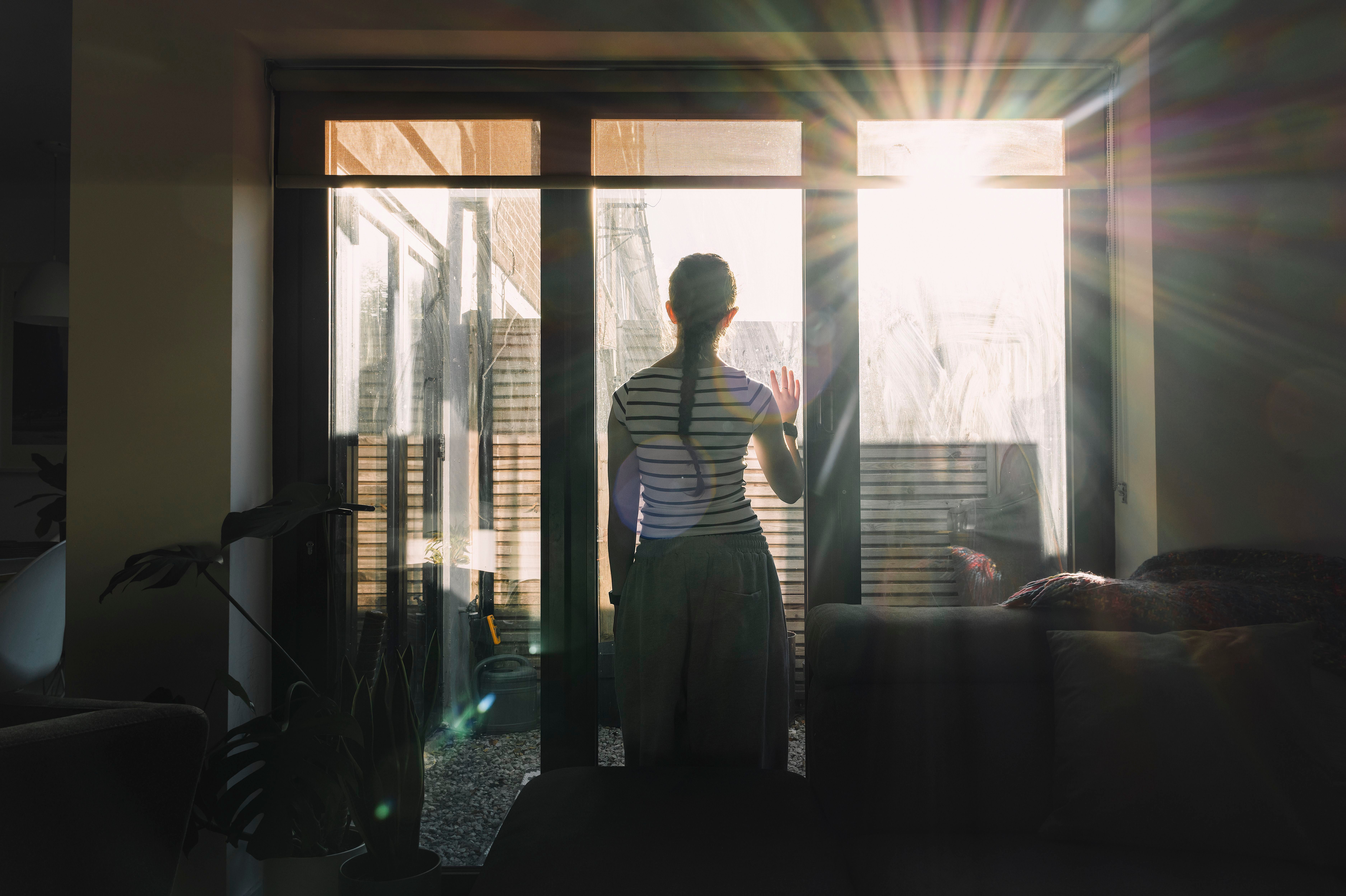 Frau steht am Fenster und blickt nach draußen in die Sonne, Sonnenstrahlen scheinen herein.