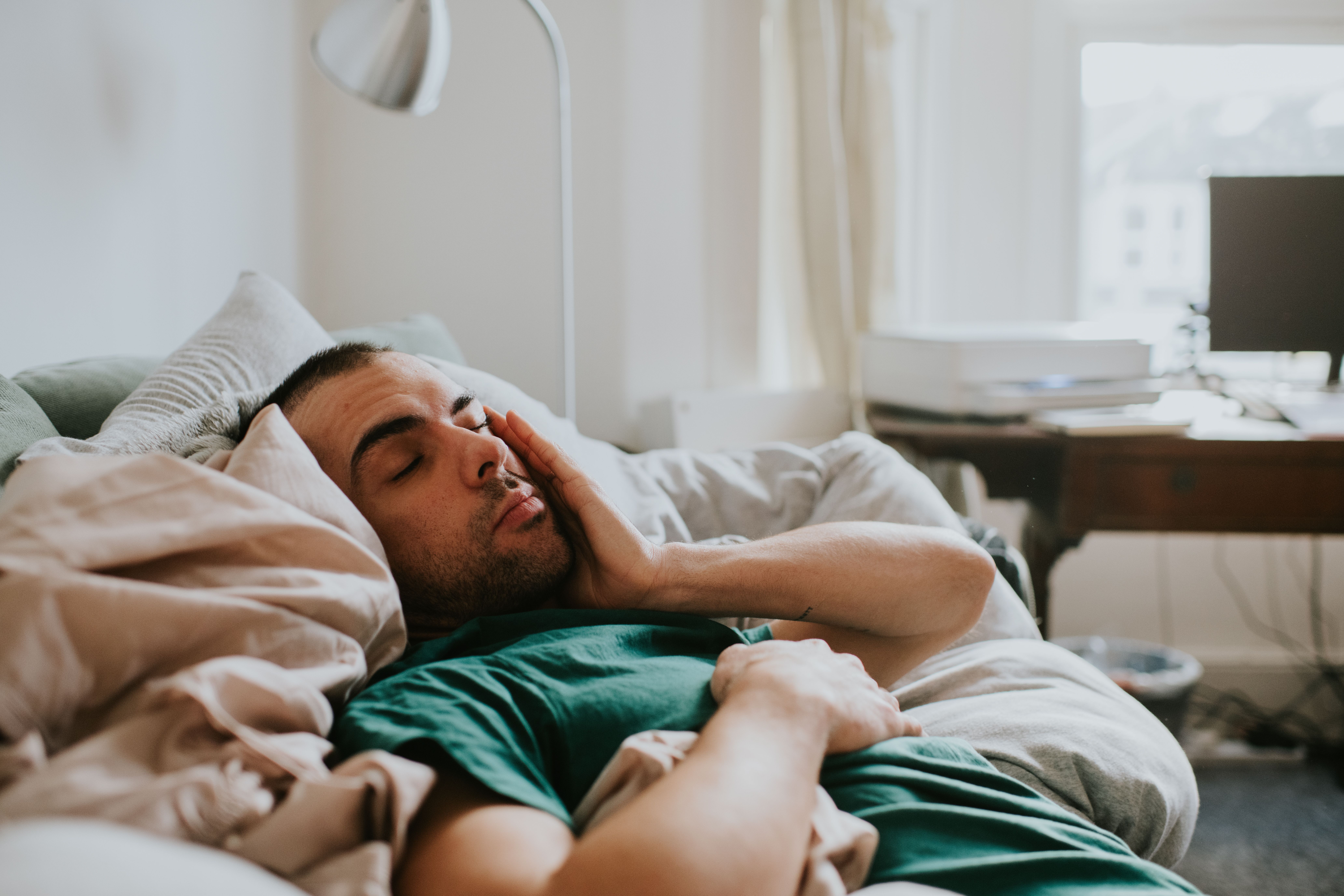 Ein Mann liegt schlaftrunken im Bett und reibt sich verschlafen das Gesicht in einem gemütlichen Zimmer.