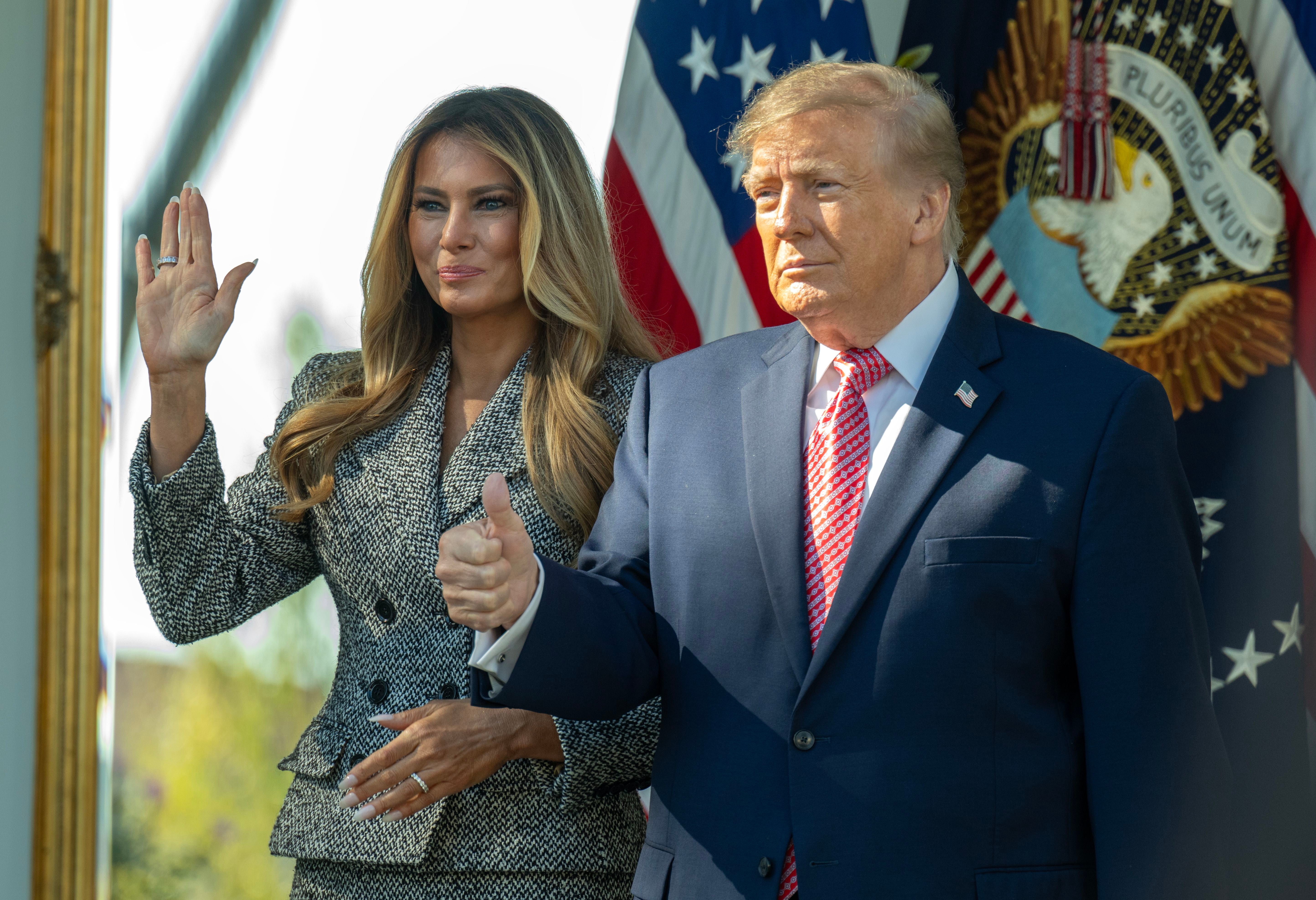 Donald Trump und Melania Trump winken und gestikulieren vor US-Flaggen im Rosengarten des Weißen Hauses.