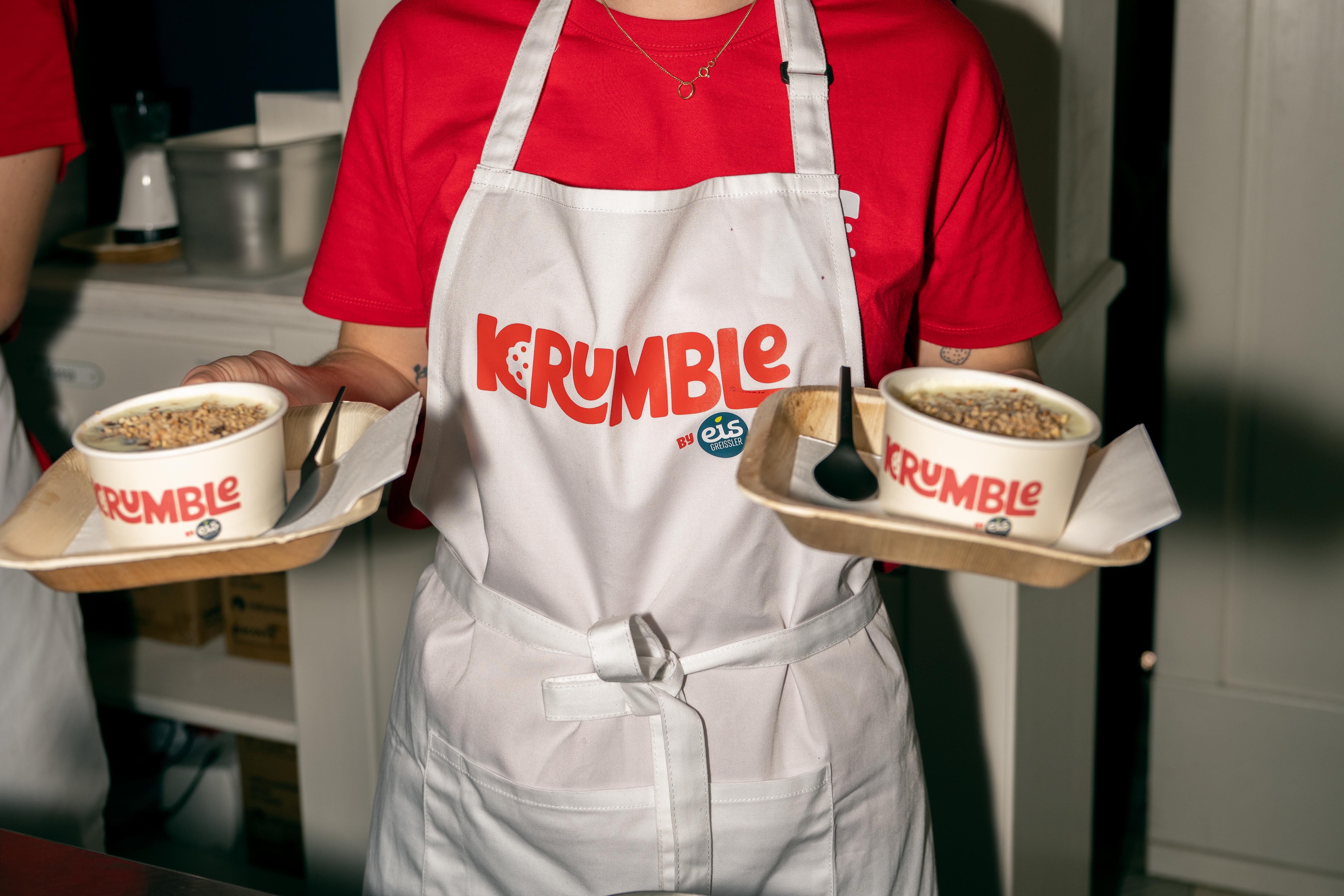 Person mit Krumble-Schürze hält zwei Tabletts mit Müslischalen und Löffeln.