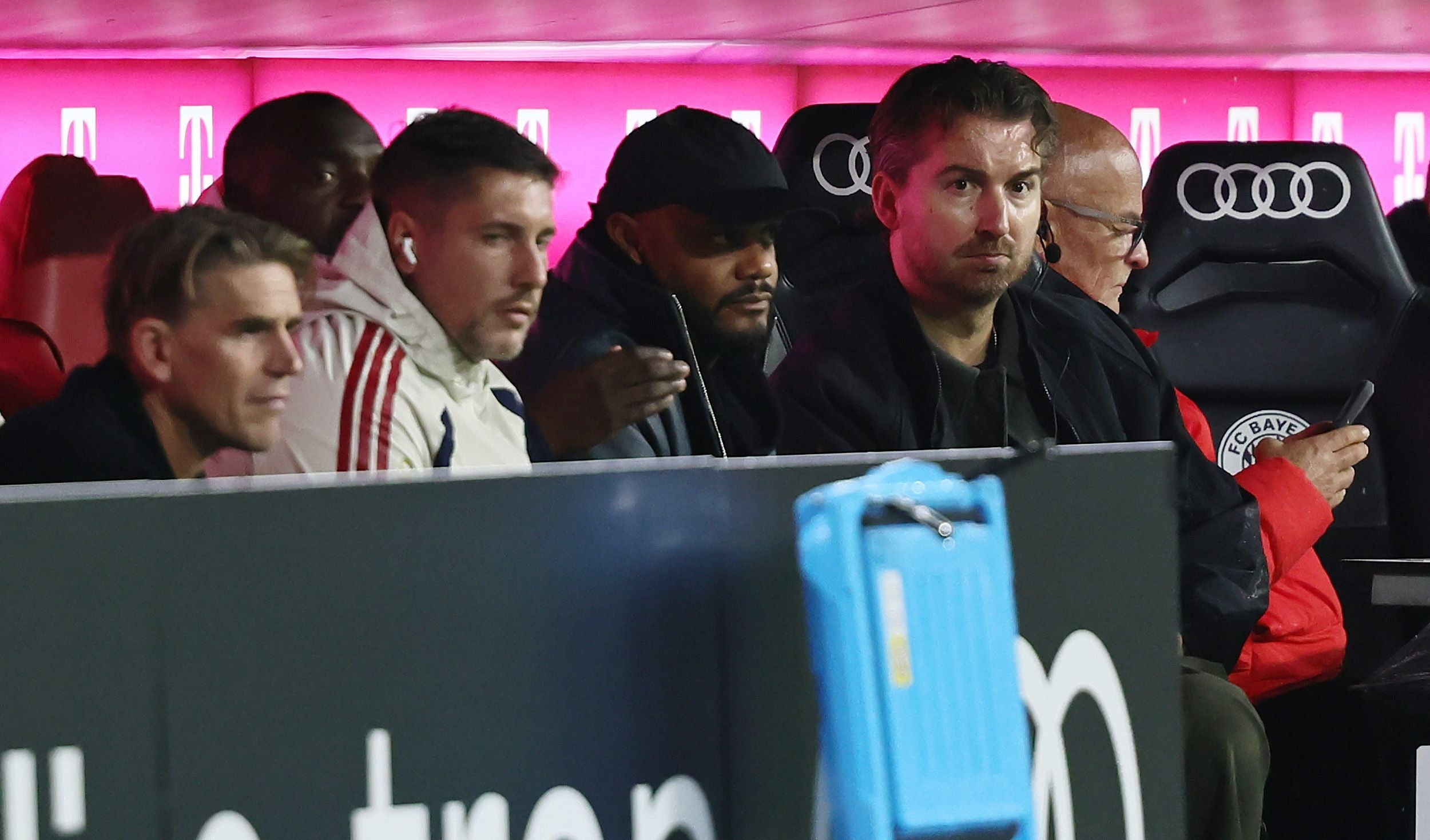 Vincent Kompany sitzt mit schwarzer Kappe und dunkler Jacke auf der Ersatzbank des FC Bayern München neben Co-Trainer Aaron Danks und Sportdirector Christoph Freund.