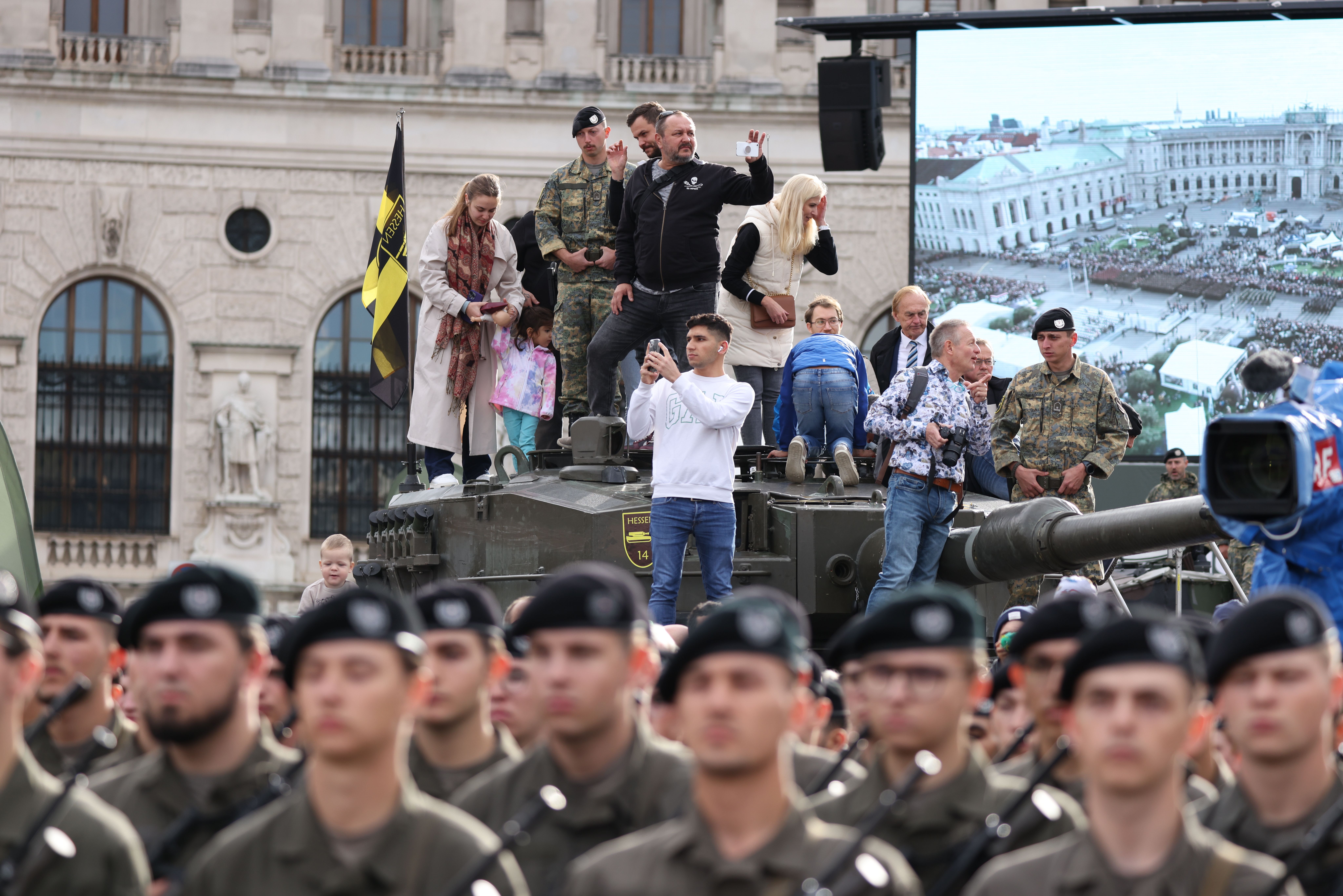 Menschen stehen auf einem Panzer, während Soldaten bei einer Angelobung am Heldenplatz marschieren.