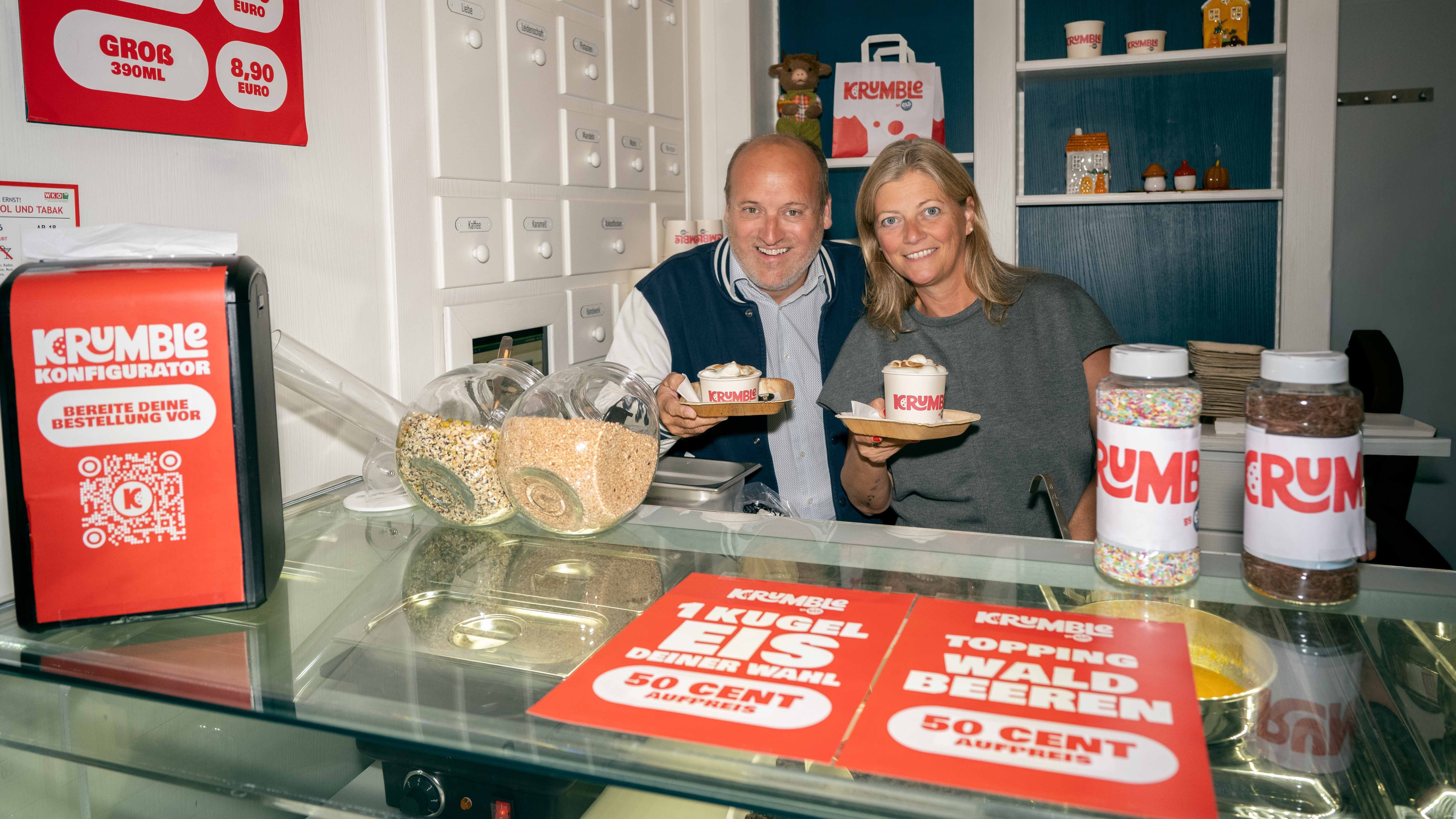Ein Mann und eine Frau halten Dessertschalen in einem Eisladen mit Toppings und Preisschildern.