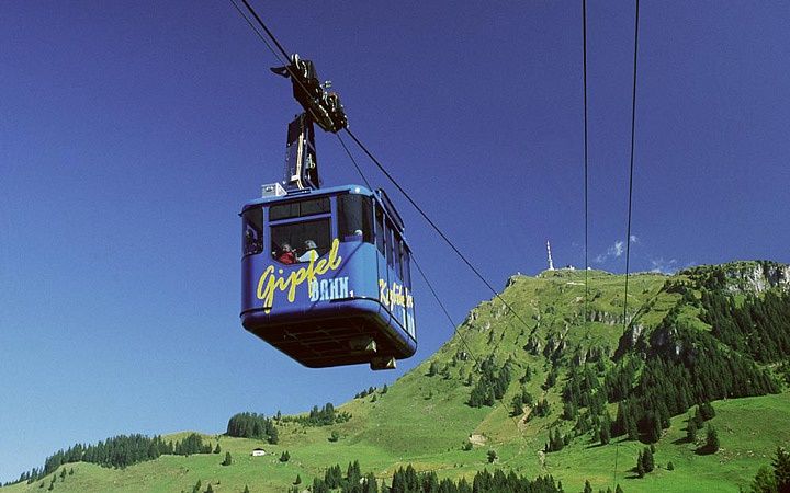 Blaue Seilbahngondel fährt vor grünem Berghang bei blauem Himmel in Kitzbühel.