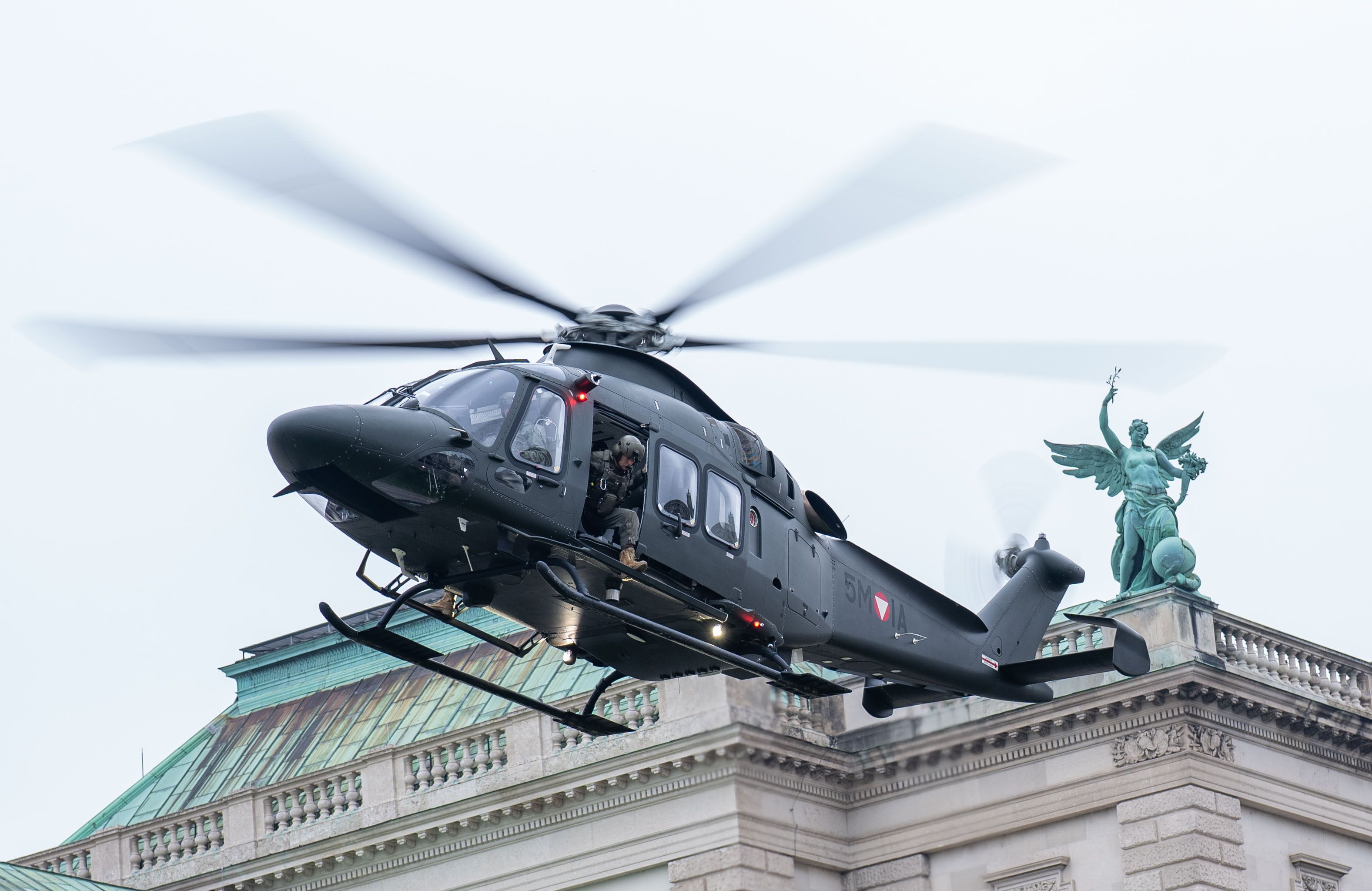 Ein schwarzer Militärhubschrauber fliegt knapp über ein historisches Gebäude am Wiener Heldenplatz.