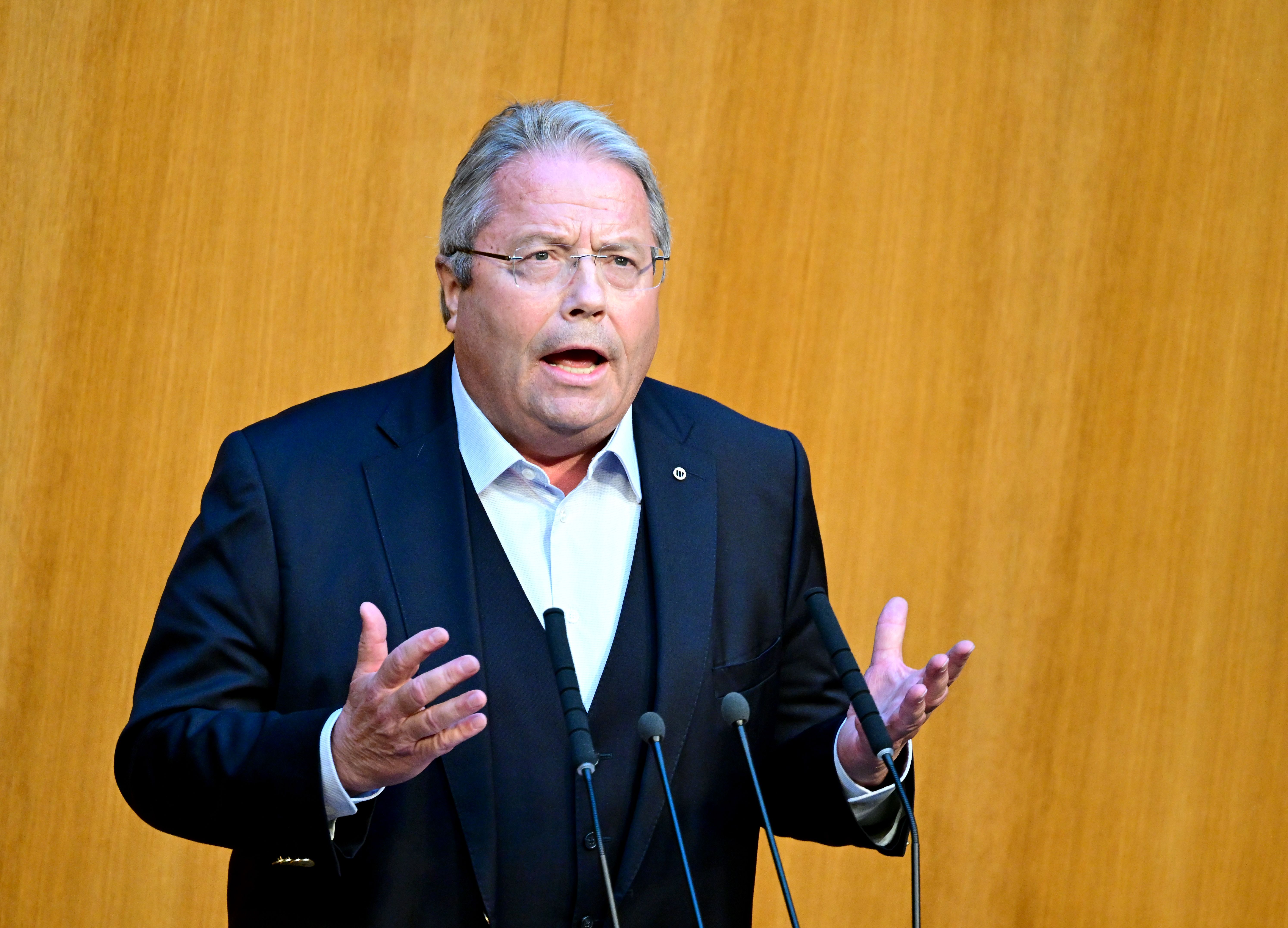 Ein Mann in Anzug spricht gestikulierend am Rednerpult vor einer holzvertäfelten Wand.