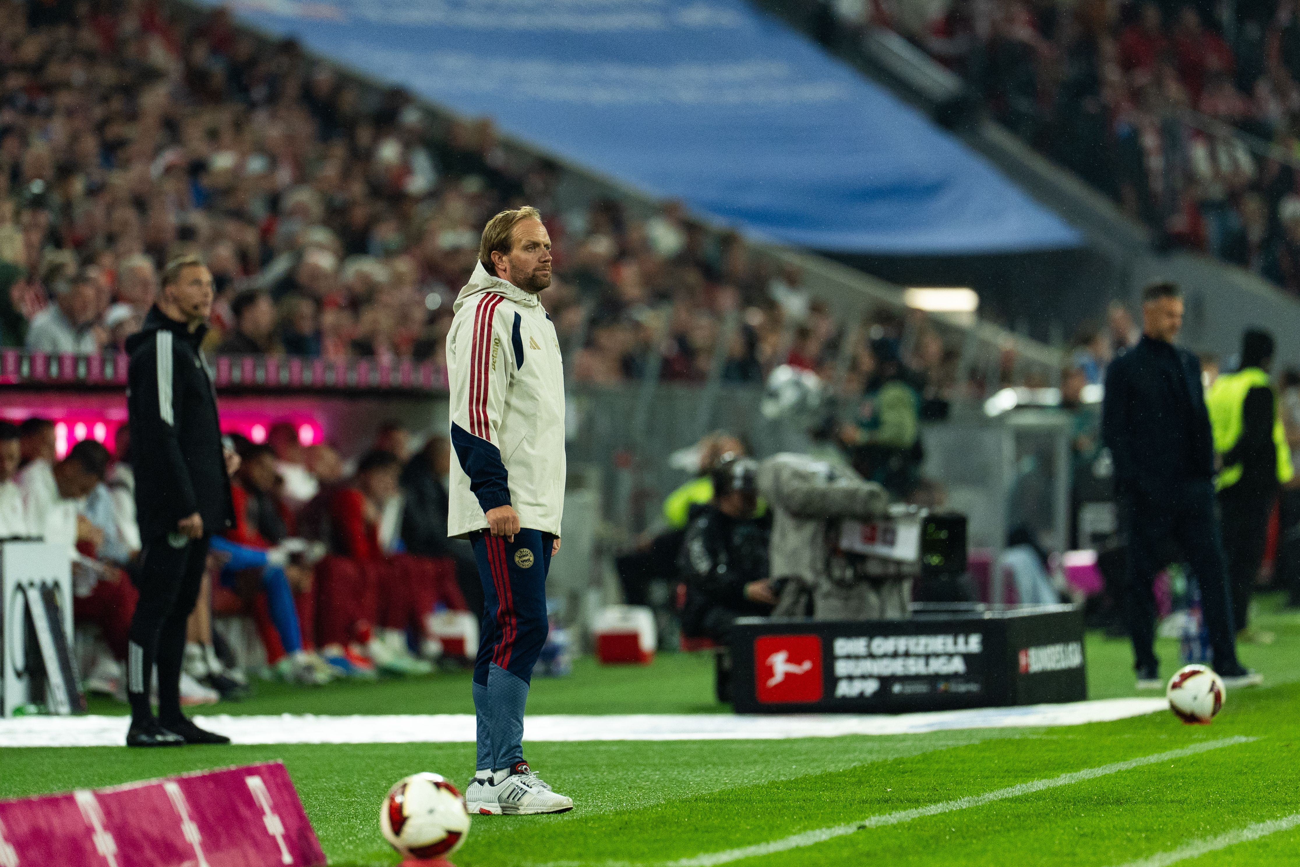 Ein Fußballtrainer gibt am Spielfeldrand während eines Spiels Anweisungen, Zuschauer im Hintergrund.