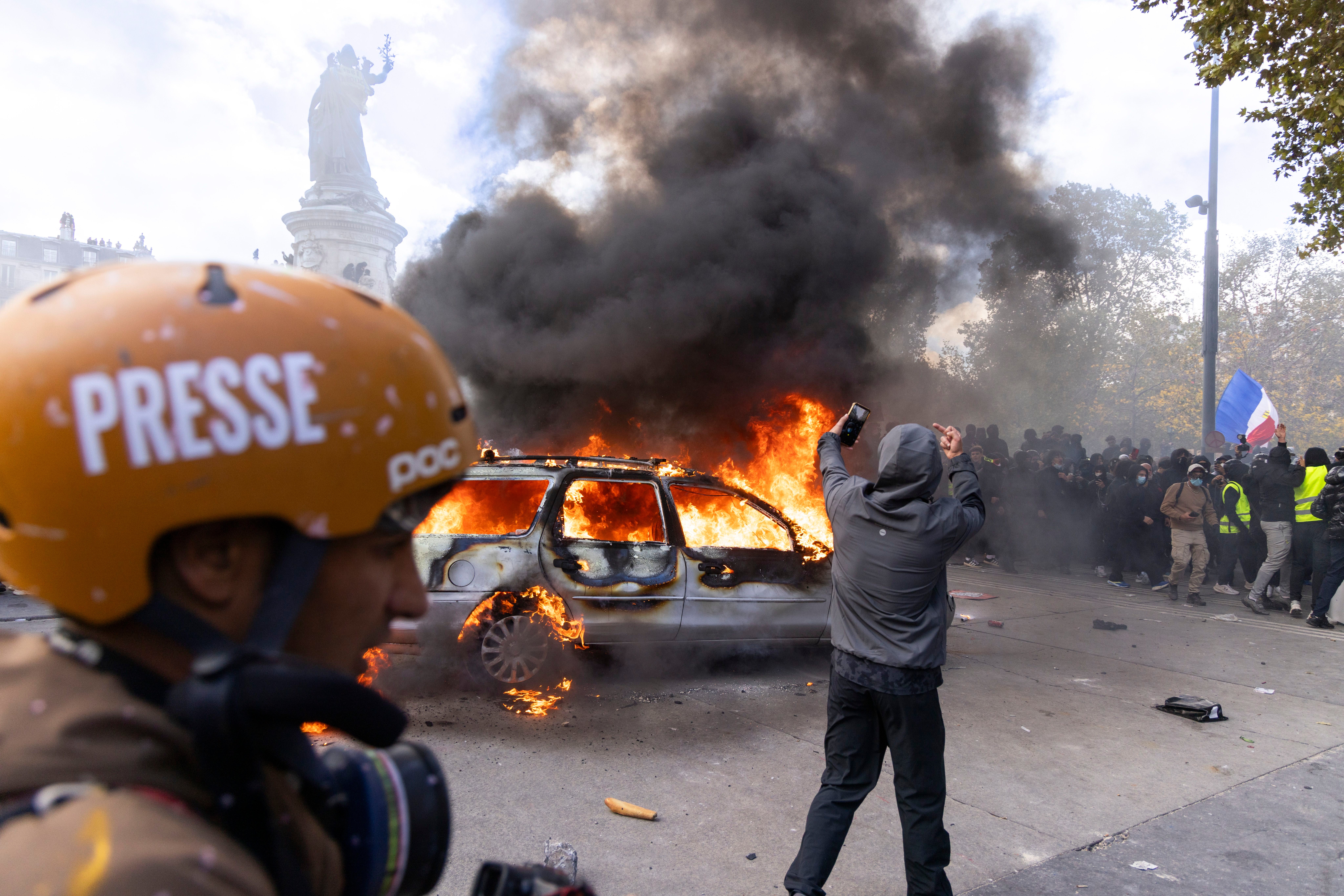 Ein Auto brennt lichterloh, dichter Rauch steigt auf, Menschen protestieren im Hintergrund.