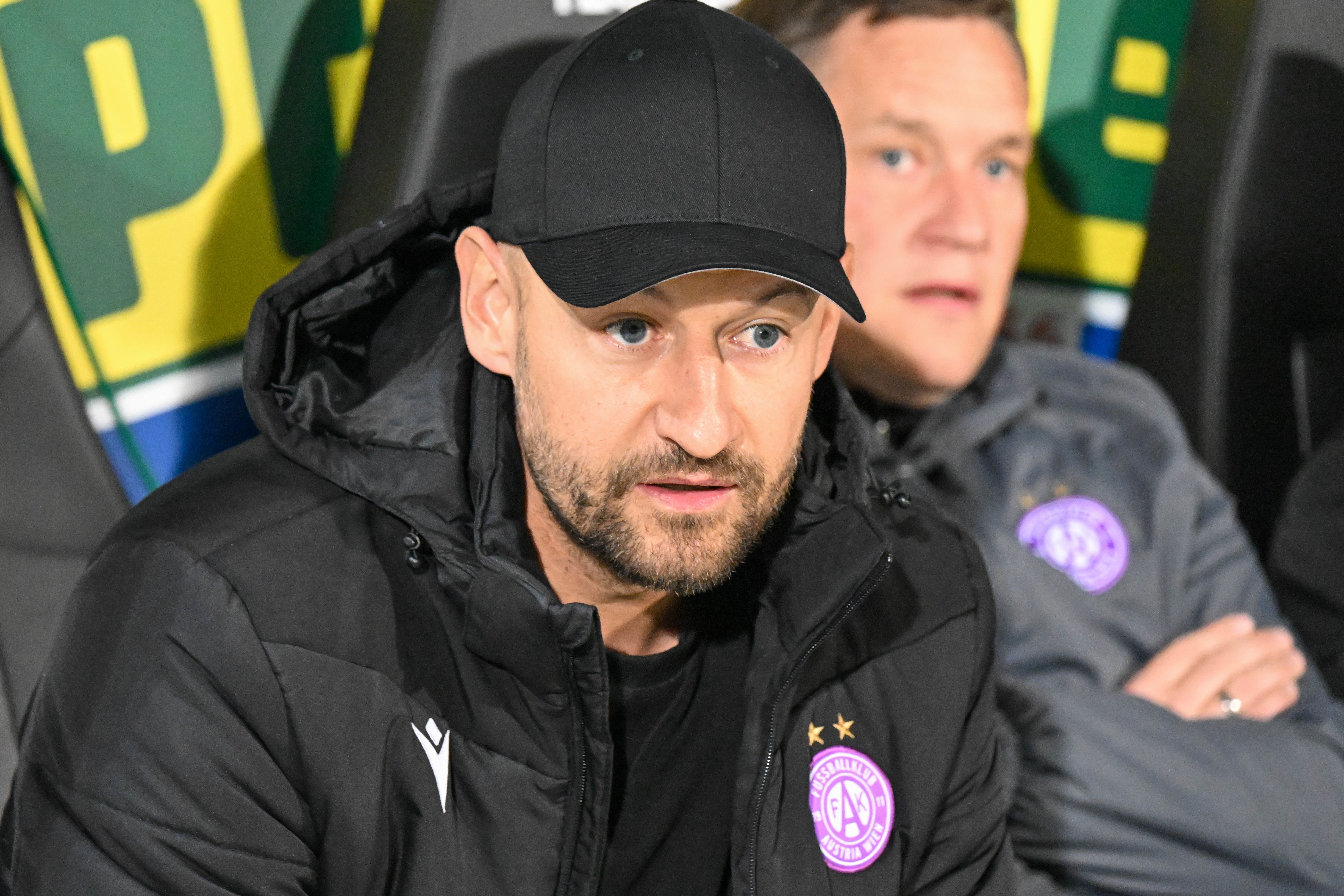 Ein Mann mit schwarzer Kappe sitzt auf einer Trainerbank, trägt Austria Wien-Jacke.