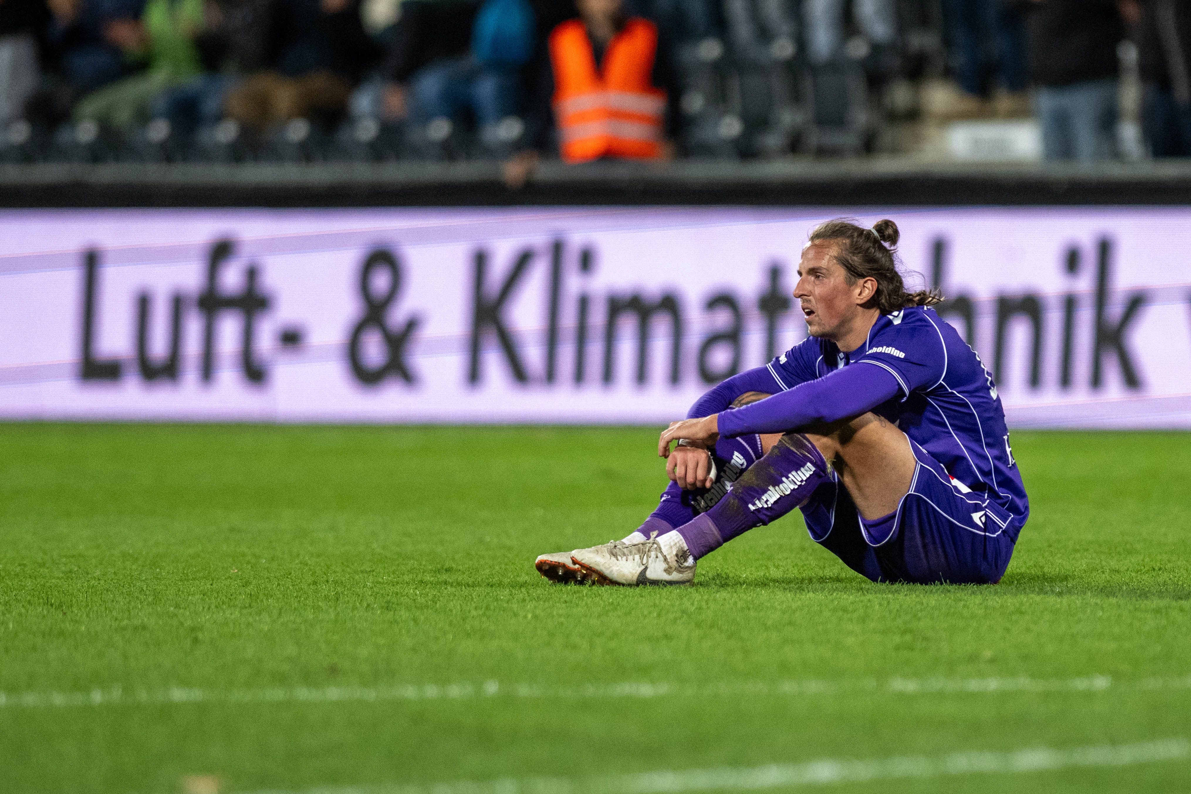 Ein Fußballspieler im lila Trikot sitzt enttäuscht auf dem Rasen eines Stadions.