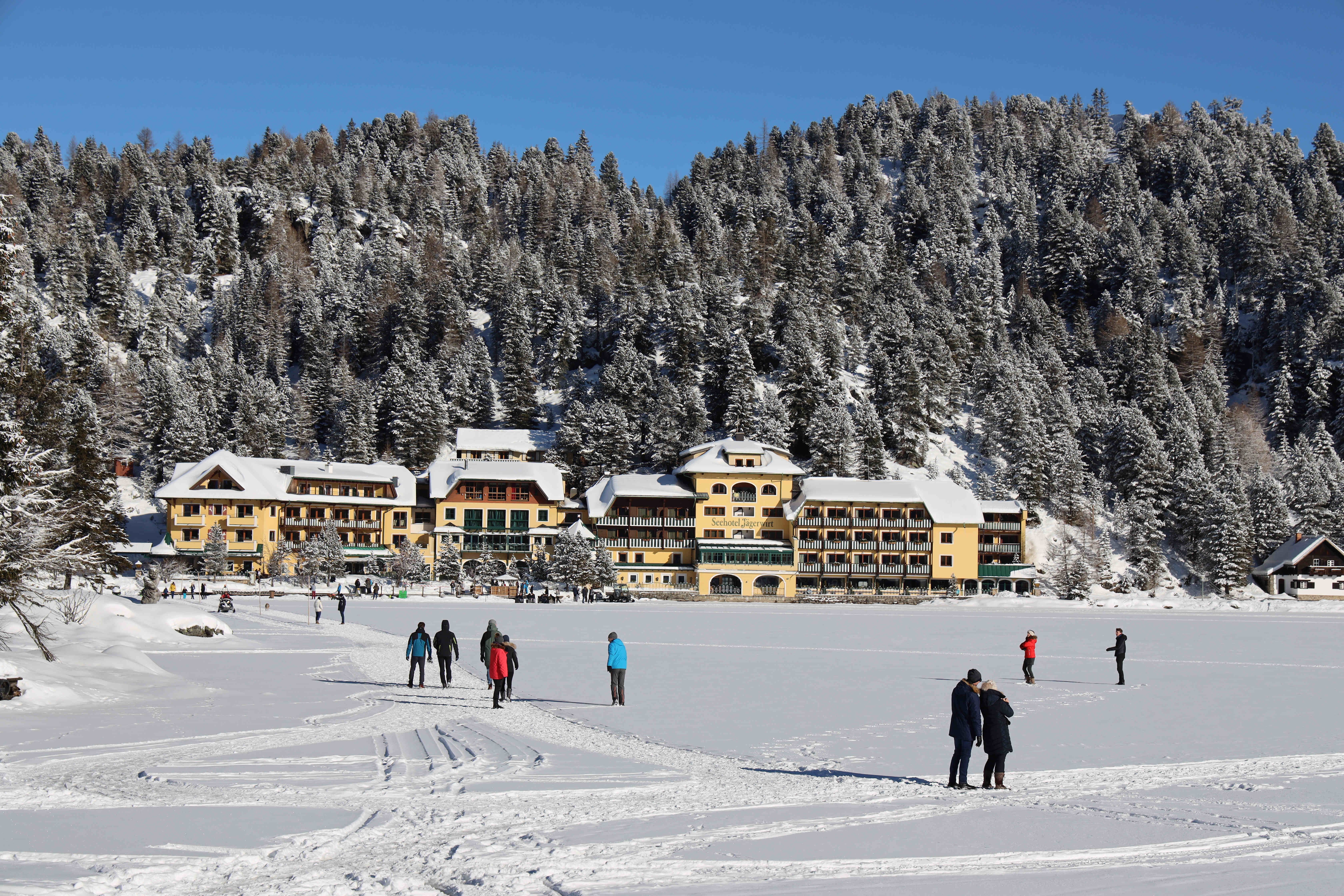 Menschen spazieren auf einem zugefrorenen See vor einem verschneiten Hotel in den Bergen.