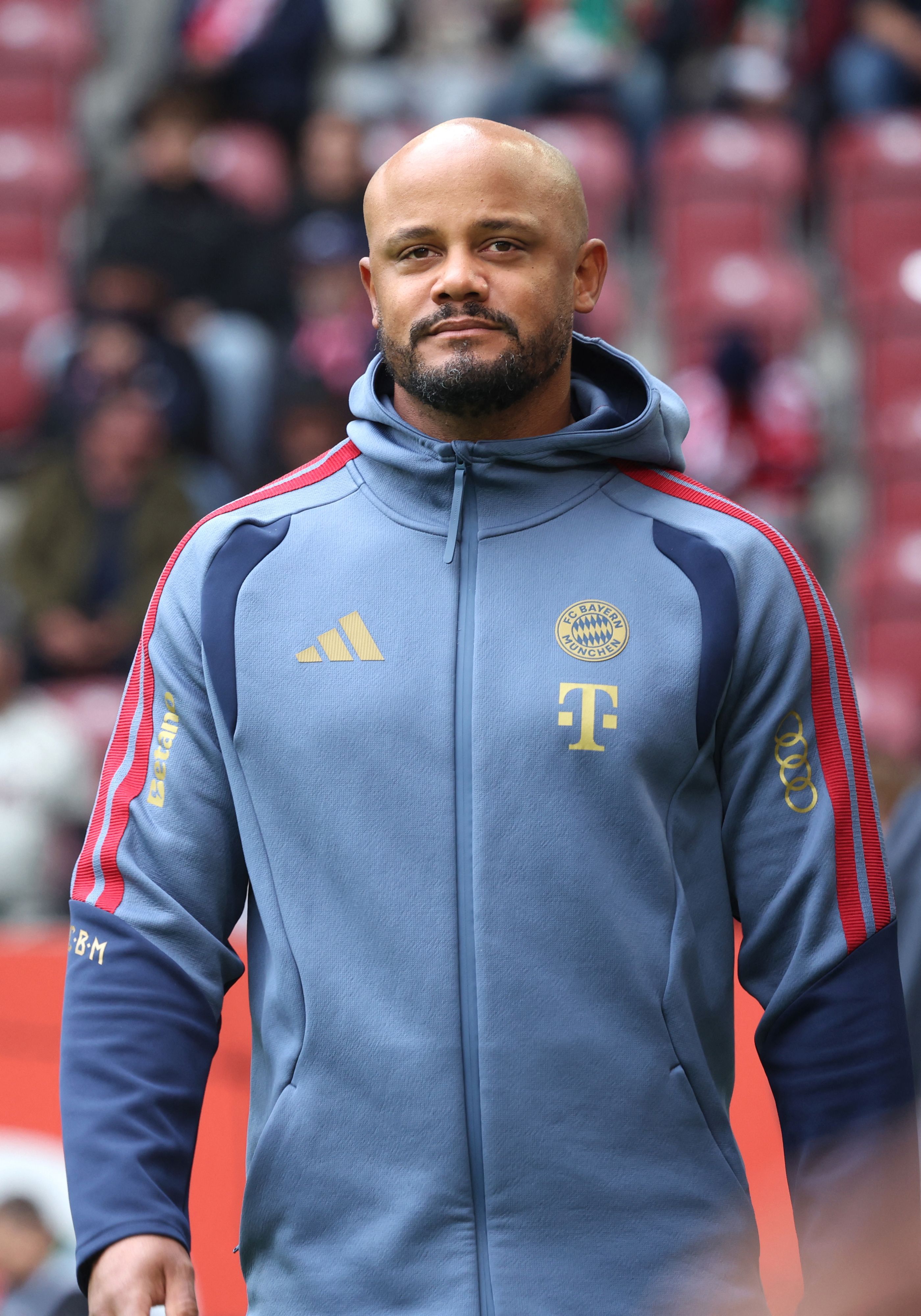 Ein Mann im Bayern München Trainingsanzug blickt ernst in die Kamera, unscharfer Stadionhintergrund.