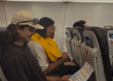 Zwei Männer sitzen im Flugzeug; einer trägt eine gelbe Rettungsweste, der andere liest die Sicherheitskarte.