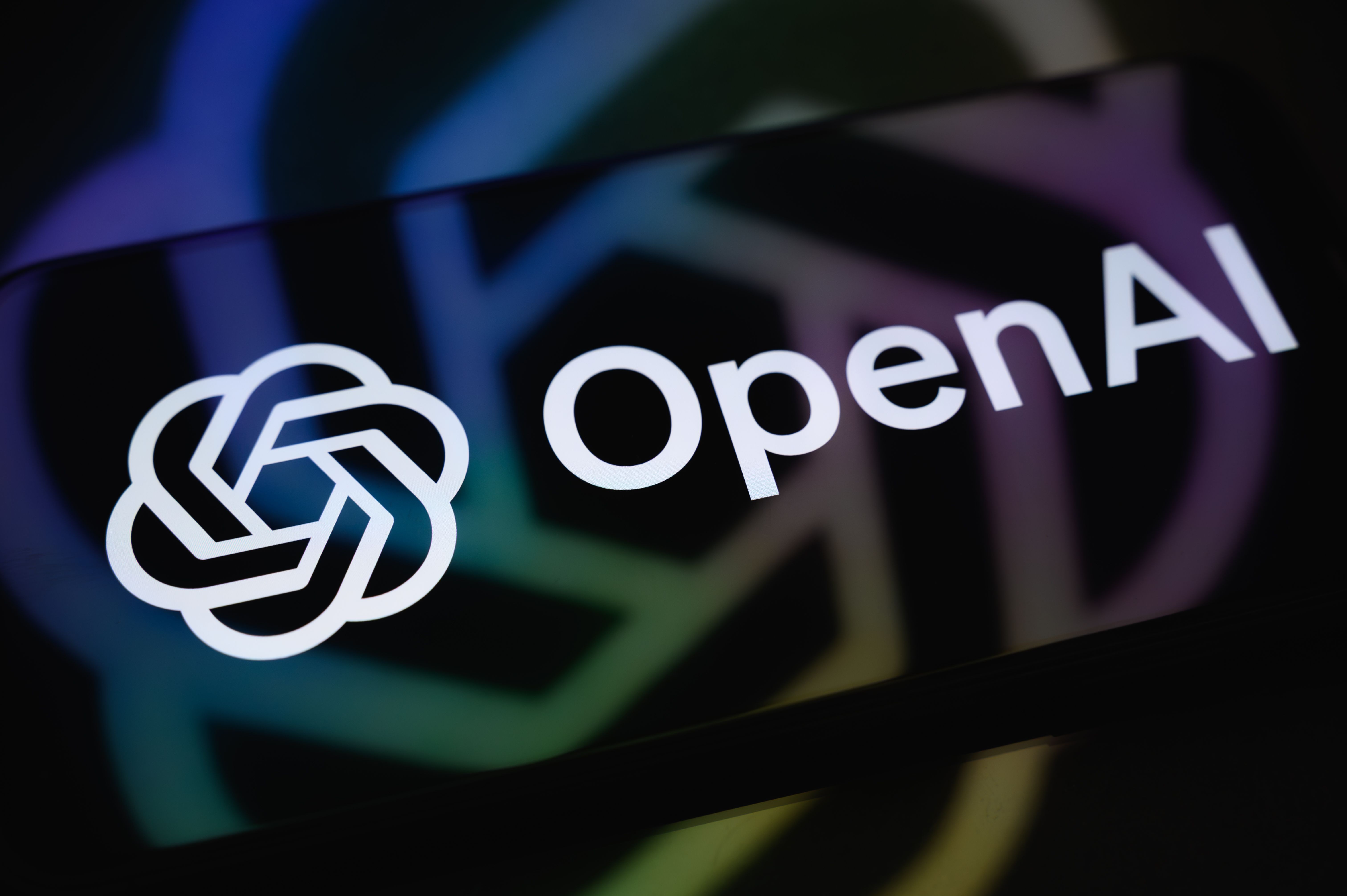 Das OpenAI-Logo und Schriftzug leuchten auf einem Smartphone mit Spiegelung im Hintergrund.
