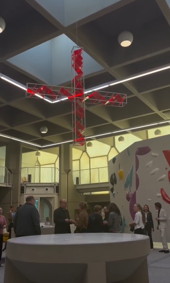Menschen stehen in einer Kirche mit Kletterwand und rotem Kreuz an der Decke.