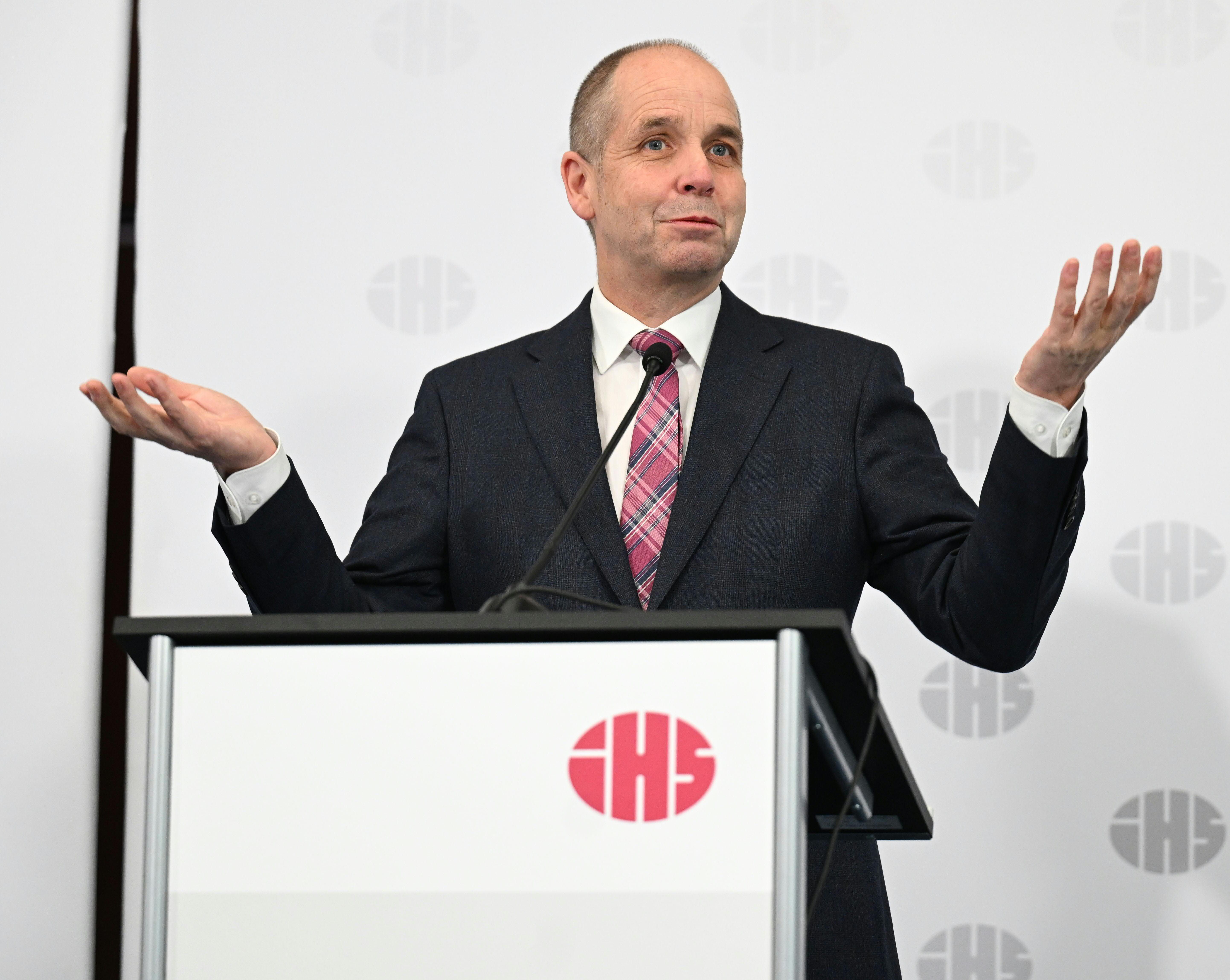 Ein Mann im Anzug spricht mit erhobenen Händen an einem Rednerpult bei einer Pressekonferenz.