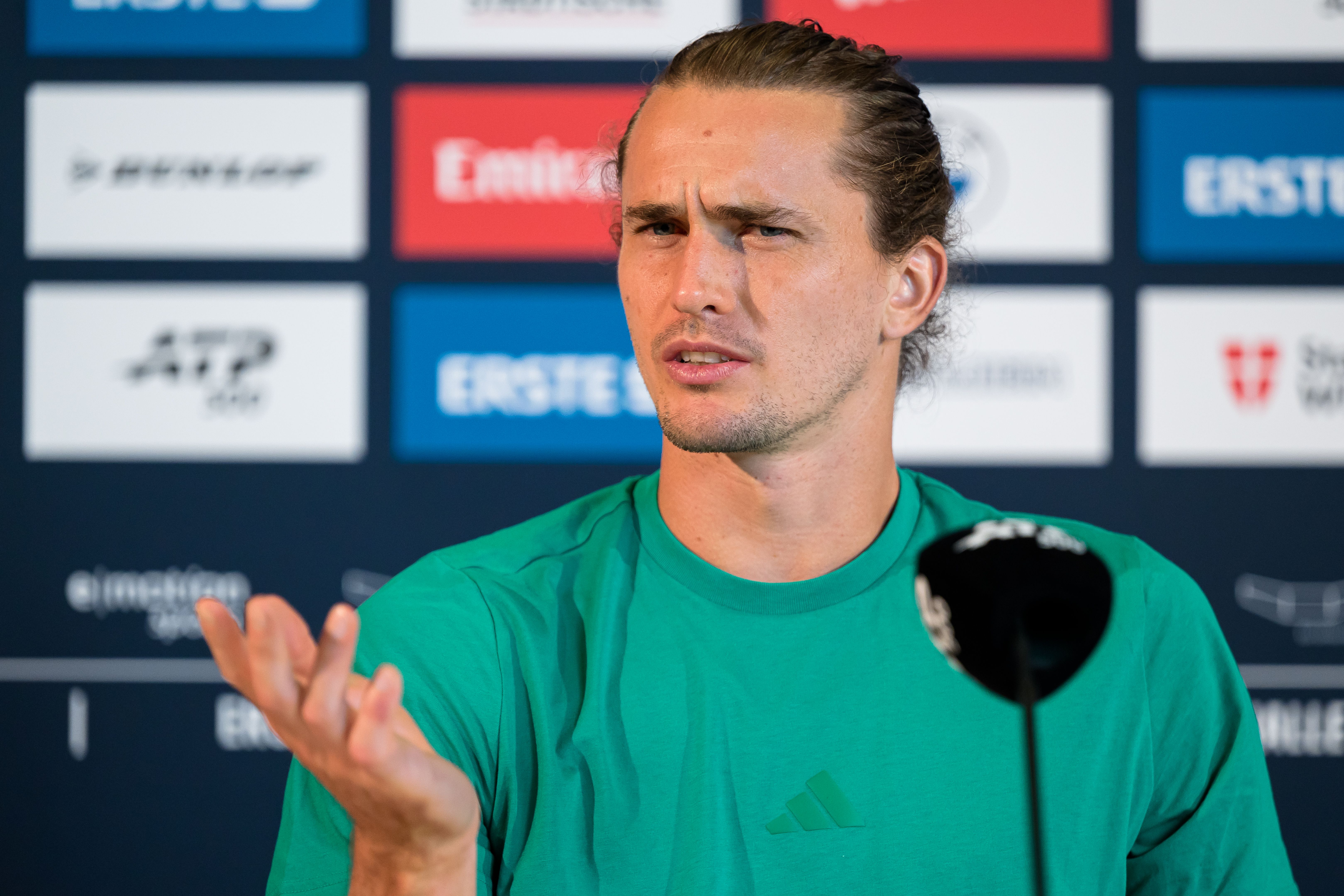 Mann in grünem Shirt gestikuliert fragend bei einer Pressekonferenz vor Sponsorenwand.